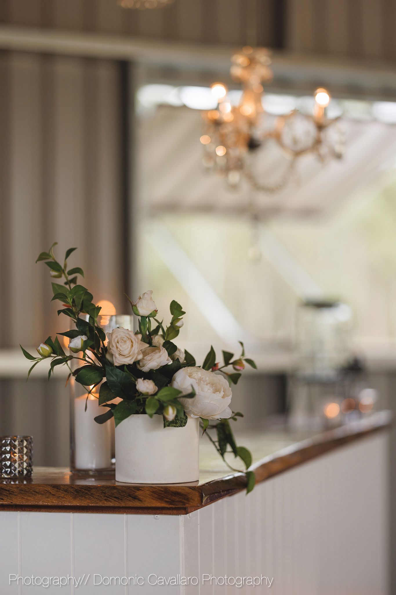 White ceramic vase with roses and greenery arrangement on wooden surface under crystal chandelier.