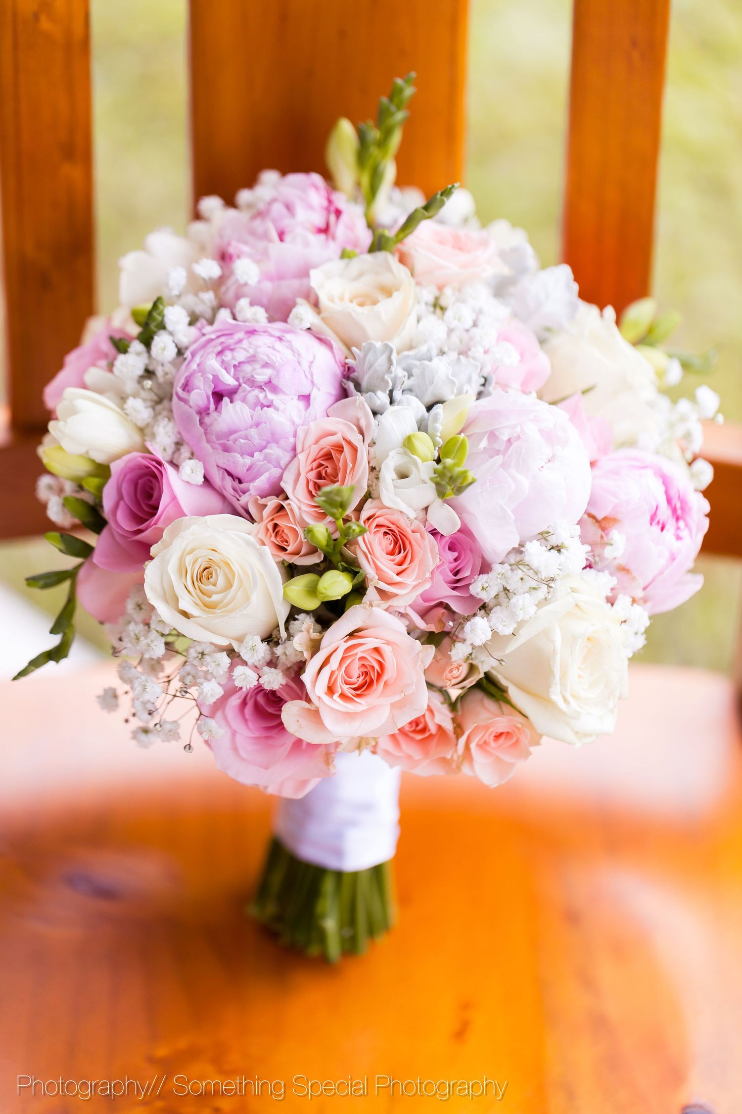 A romantic wedding bouquet featuring pink peonies, peach roses and white flowers tied with a white ribbon against a wooden backdrop.