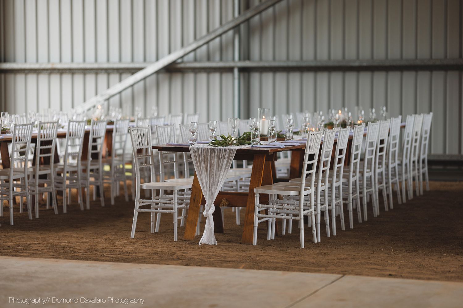 Long rows of white wooden chairs and rustic wooden tables set up in a barn for a wedding reception or event.