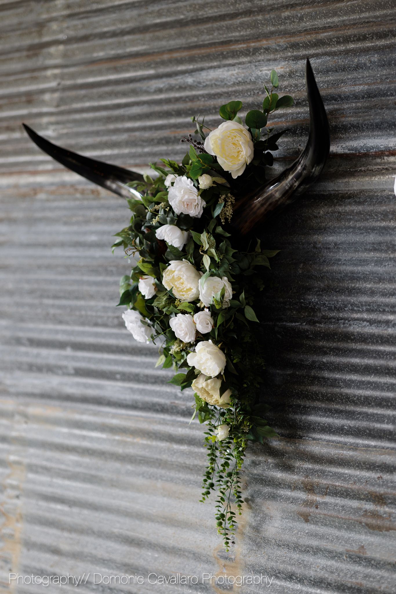 Decorative deer antlers adorned with white roses and cascading greenery against a corrugated metal wall.