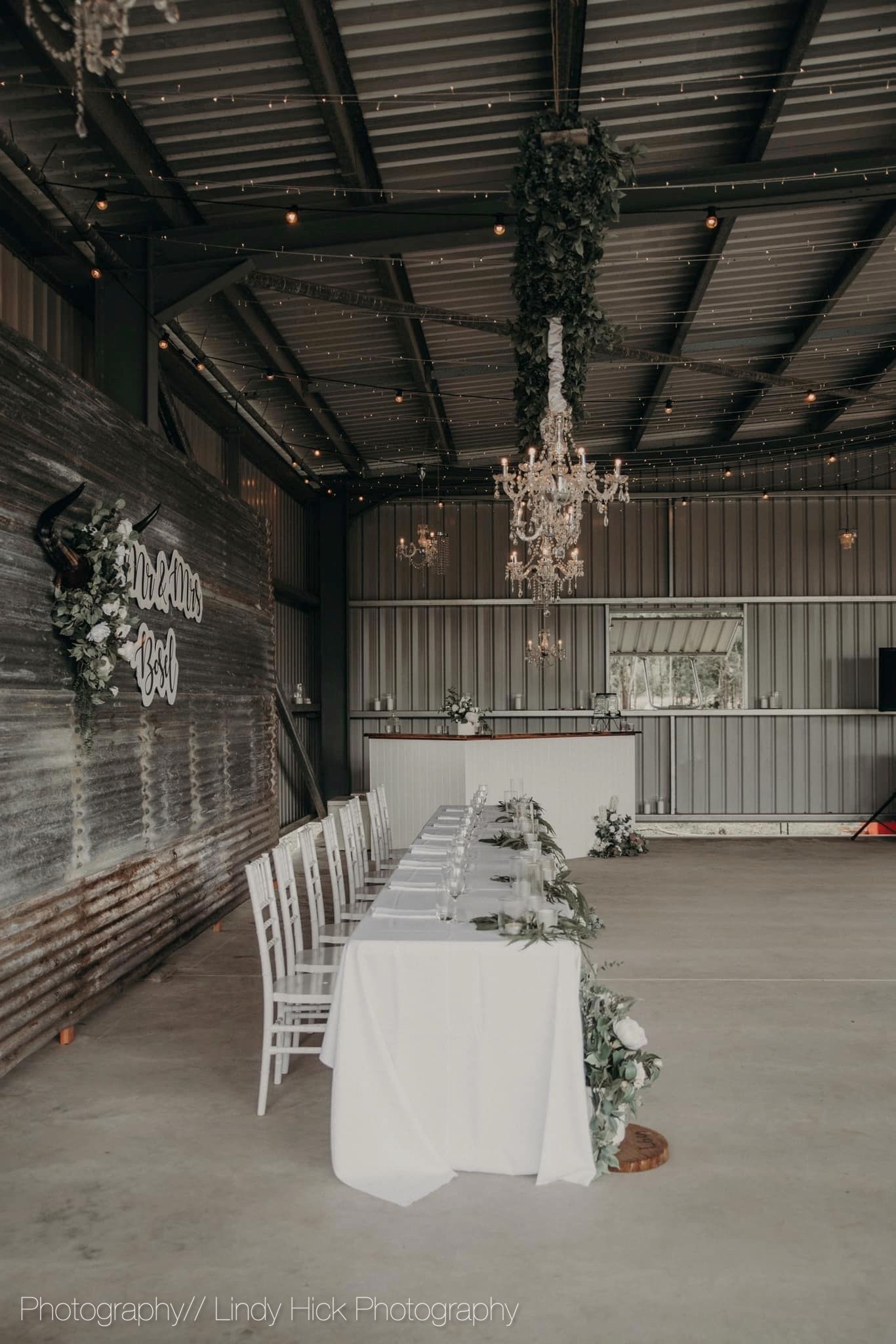 Rustic wedding reception venue with white table, chairs and elegant chandelier in an industrial barn setting.