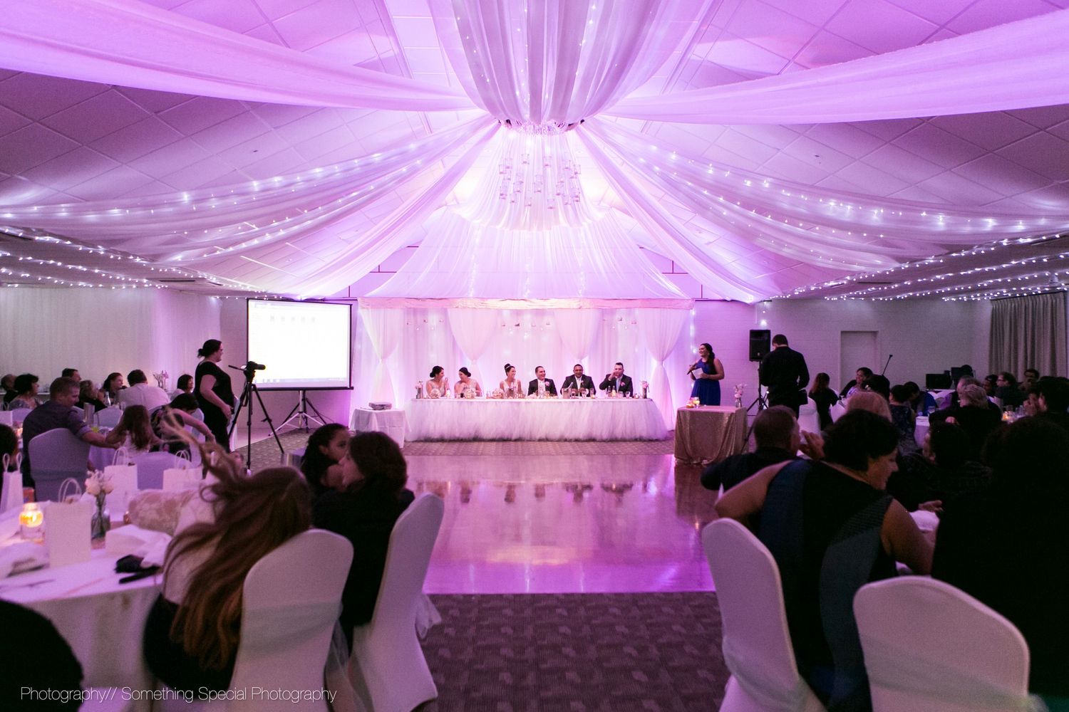 Wedding reception venue with pink-purple uplighting and draped ceiling in a white tent featuring a head table and guests seated.