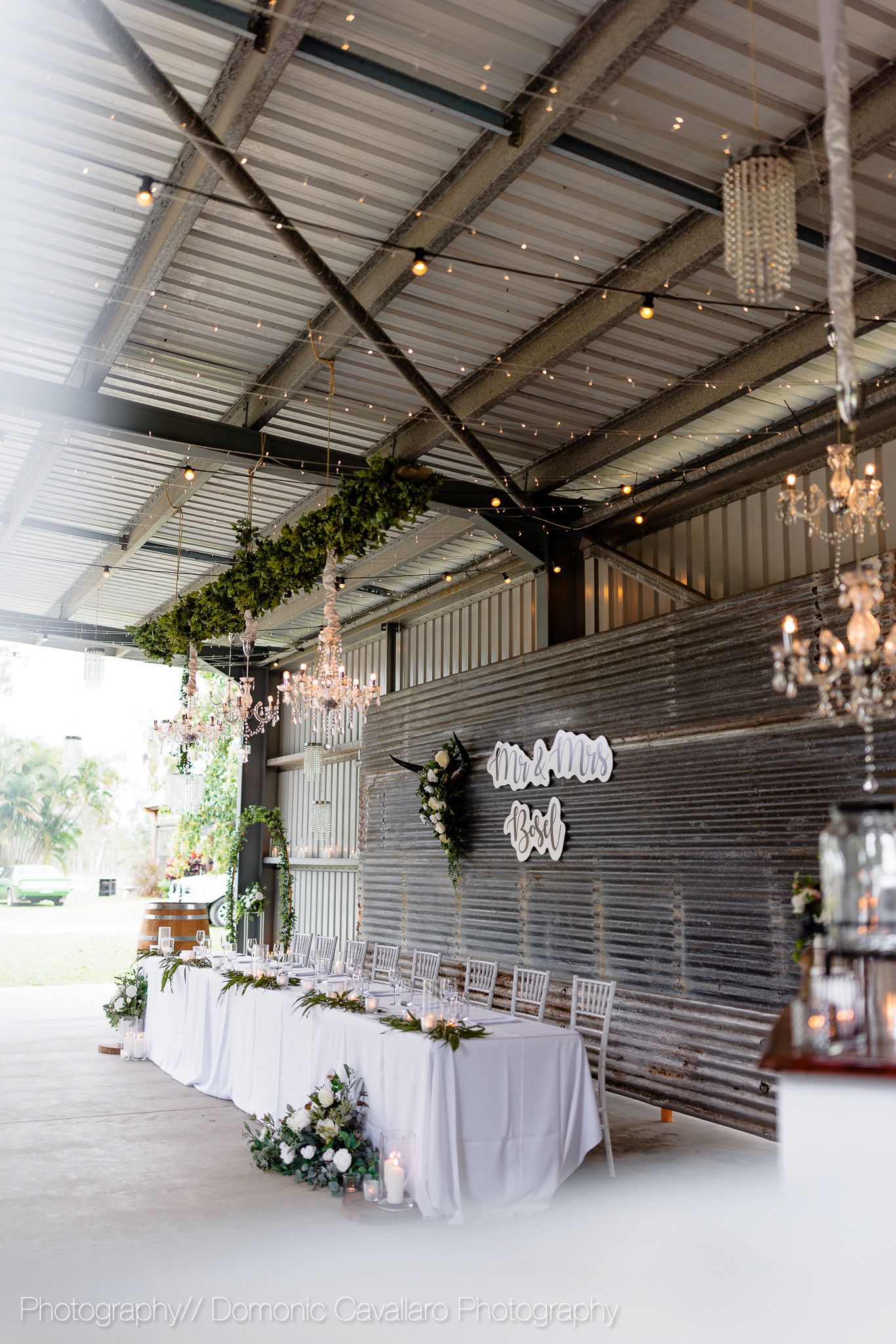 Rustic barn wedding reception setup with chandeliers, string lights, and white tablecloths along metal walls.