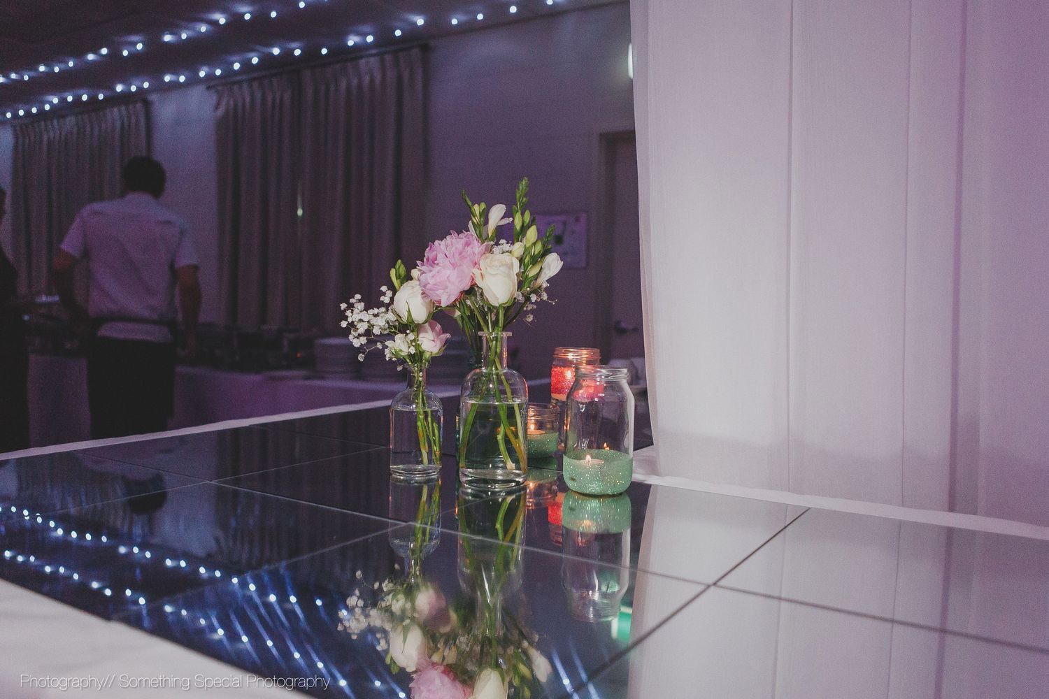 Floral centerpieces in glass vases reflect on a glossy table surface with blue string lights glowing in the background.
