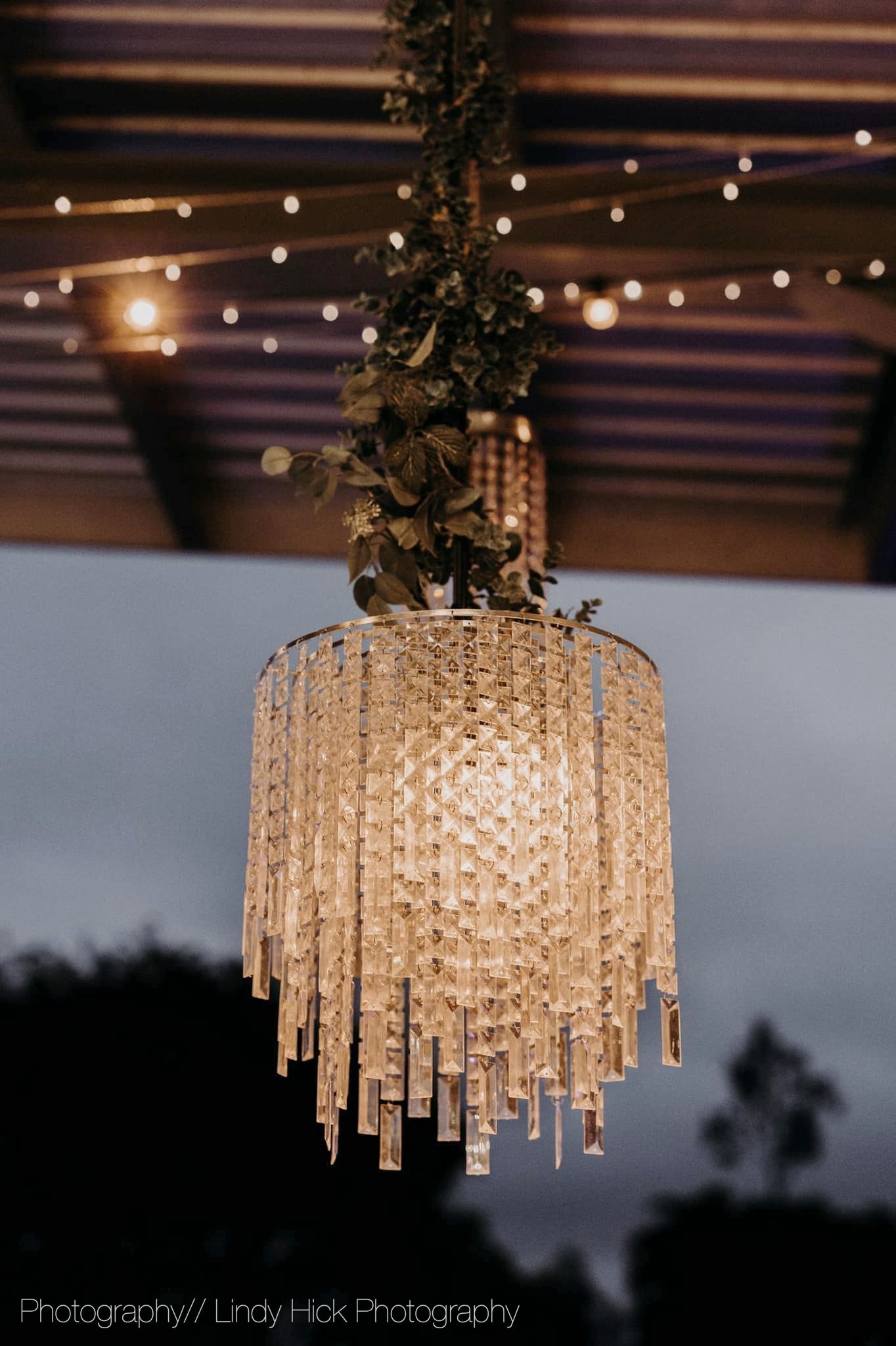 Elegant crystal chandelier with cascading glass prisms hanging from rustic ceiling with string lights and greenery.