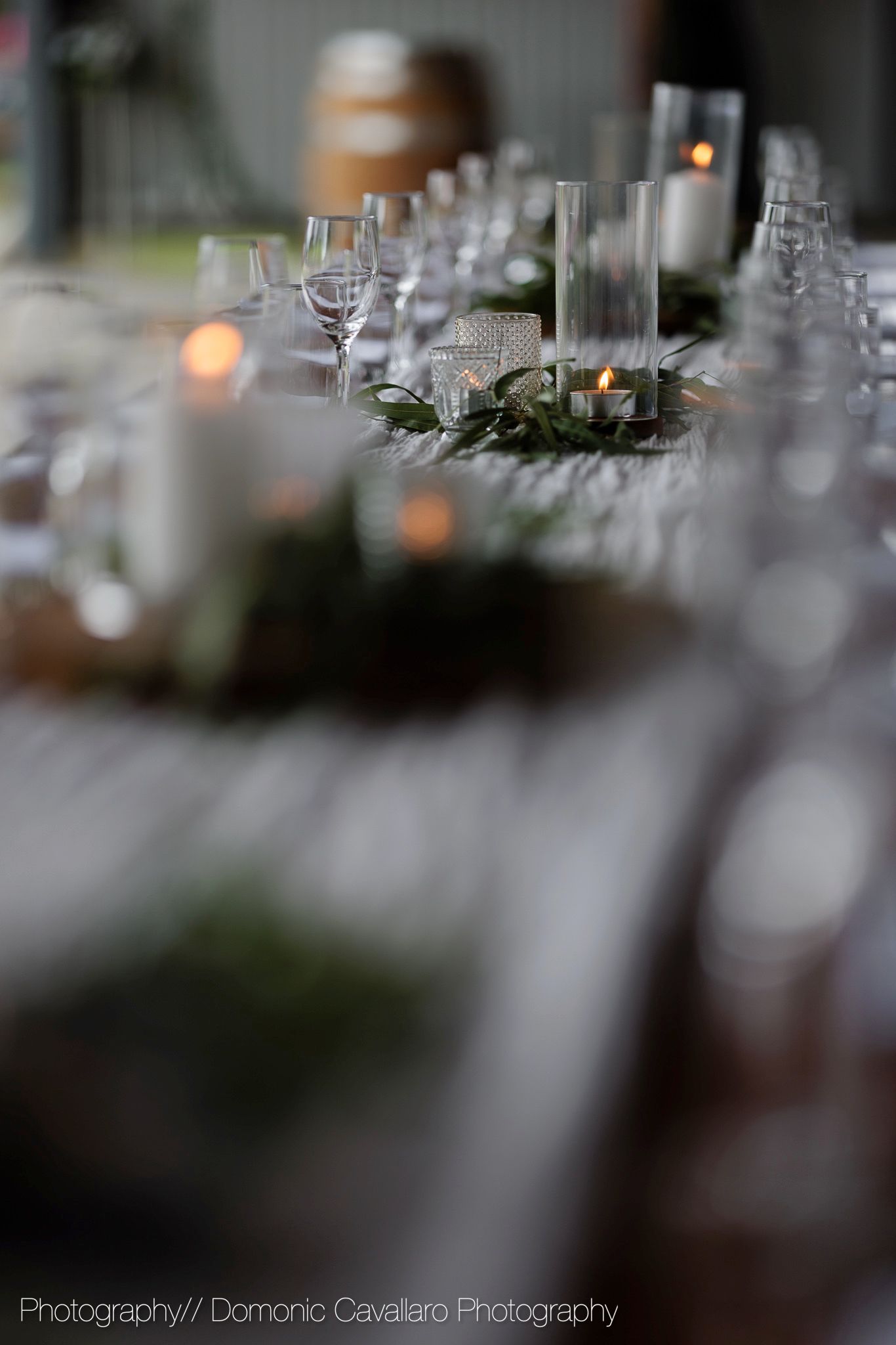 Elegant table setting at wedding reception with glowing candles and wine glasses arranged along white tablecloth.