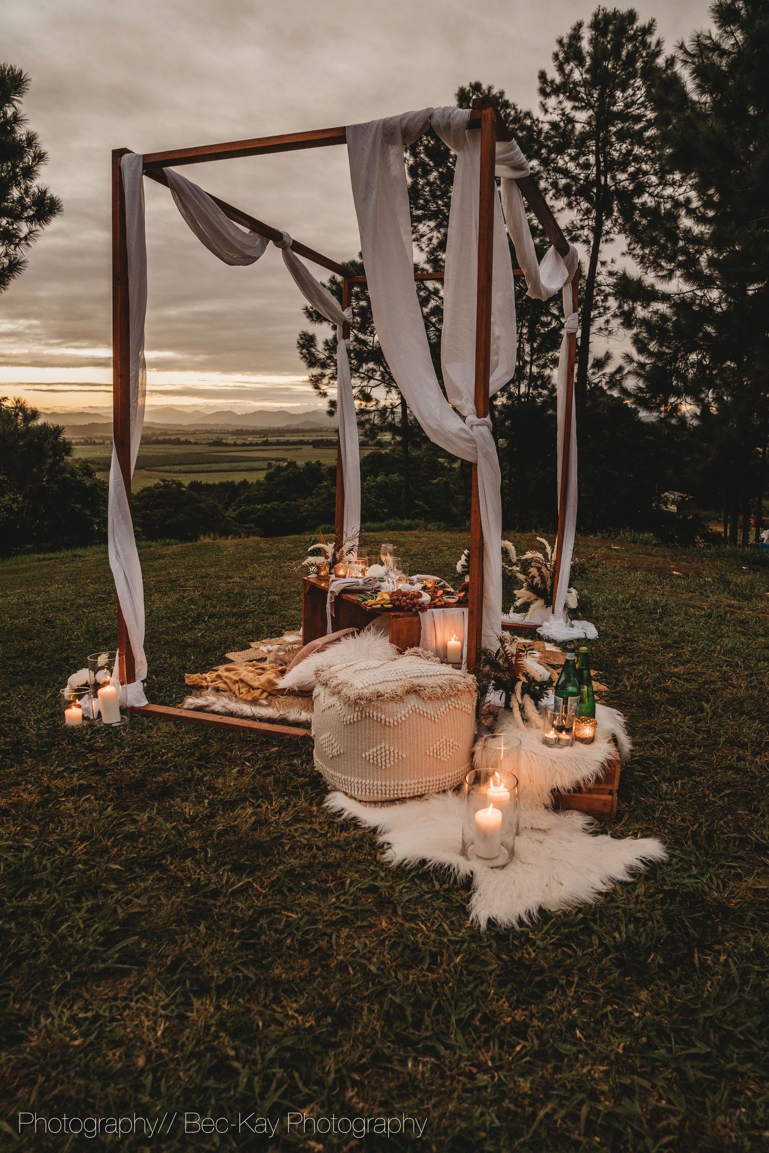 A romantic outdoor picnic setup at sunset with wooden pergola, draped fabric, fur rugs, lanterns and scattered roses.