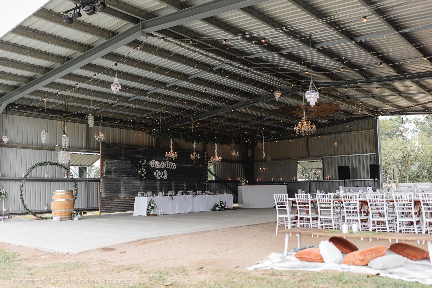 Open-air barn venue with rustic chandeliers and white chairs set up for wedding reception.