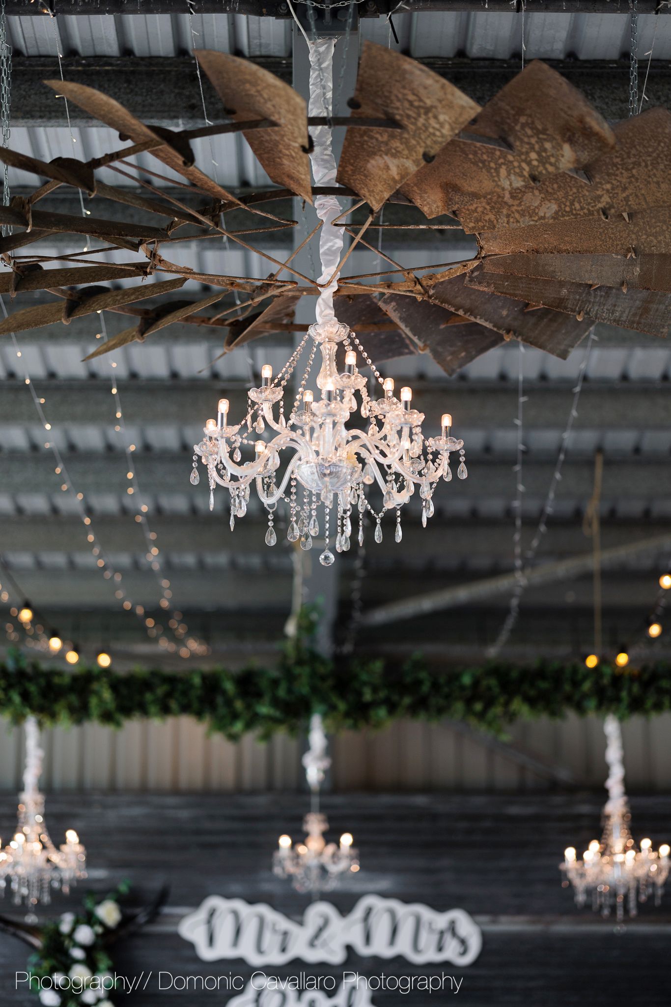 Crystal chandelier with multiple tiers hangs elegantly from a rustic barn ceiling with fairy lights and greenery.