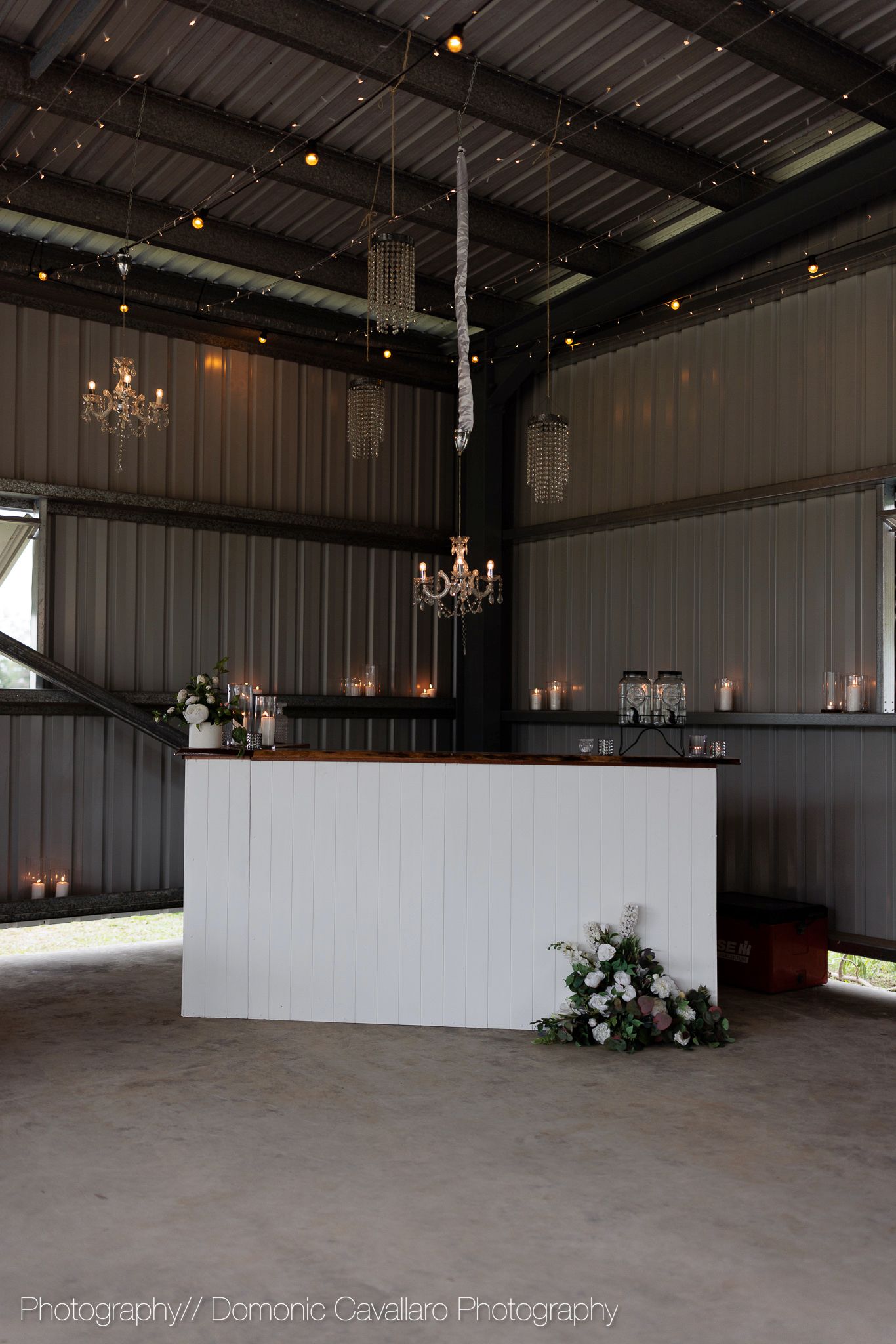 Modern rustic wedding bar setup with crystal chandeliers and white counter decorated with floral arrangements.