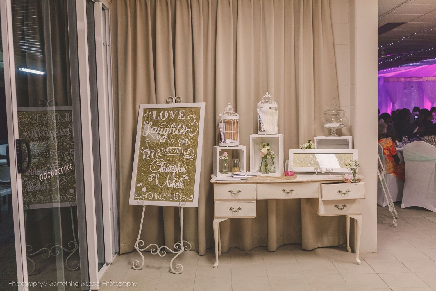 Vintage white vanity desk with wedding photos and chalkboard sign displayed against beige curtain at reception venue.