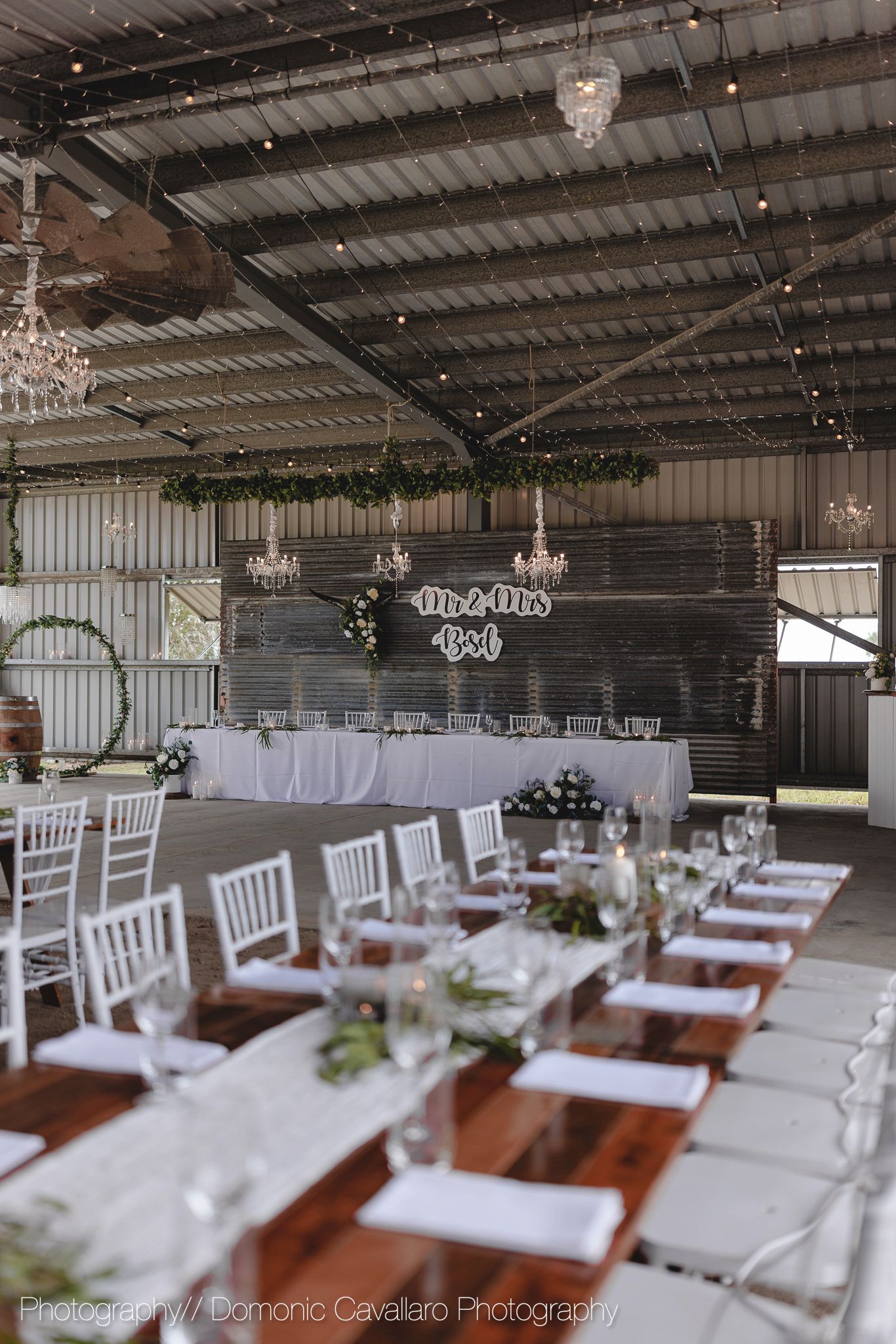Rustic barn wedding reception setup with wooden tables, white chairs, and crystal chandeliers hanging from metal ceiling.