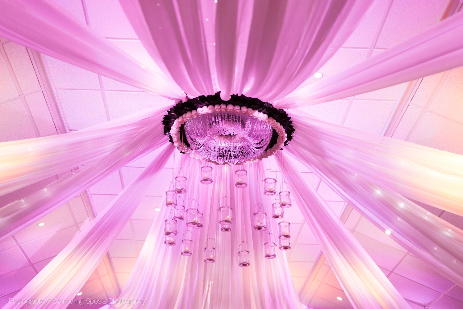 Elegant circular chandelier with draping white fabric and pink uplighting creating a romantic wedding reception atmosphere.