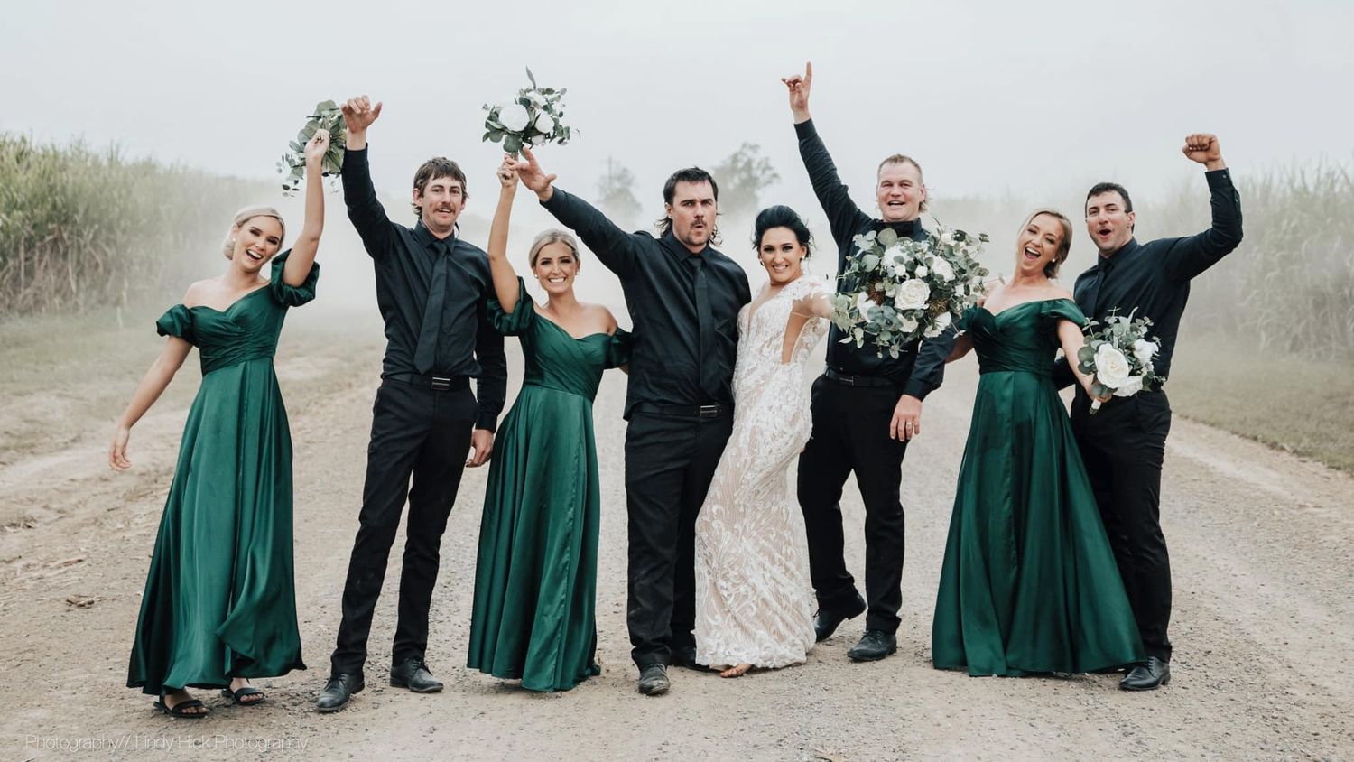 A wedding party celebrating on a misty path with bridesmaids in emerald green dresses and groomsmen in black suits.
