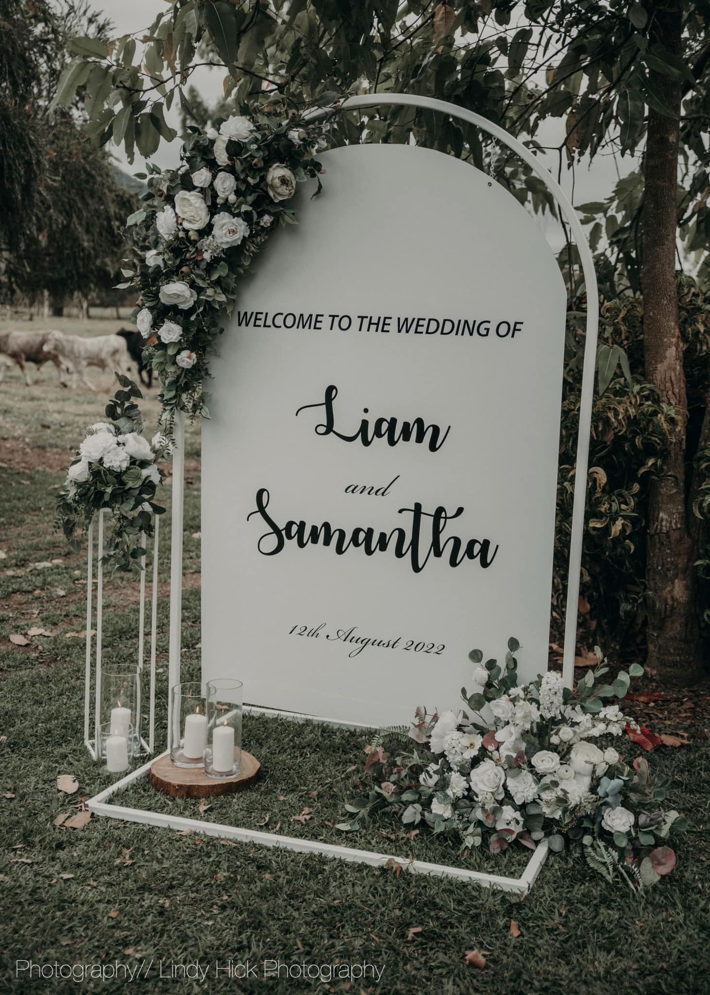 Elegant white wedding welcome sign with floral arch displays 'Liam and Samantha' in script lettering with white roses.