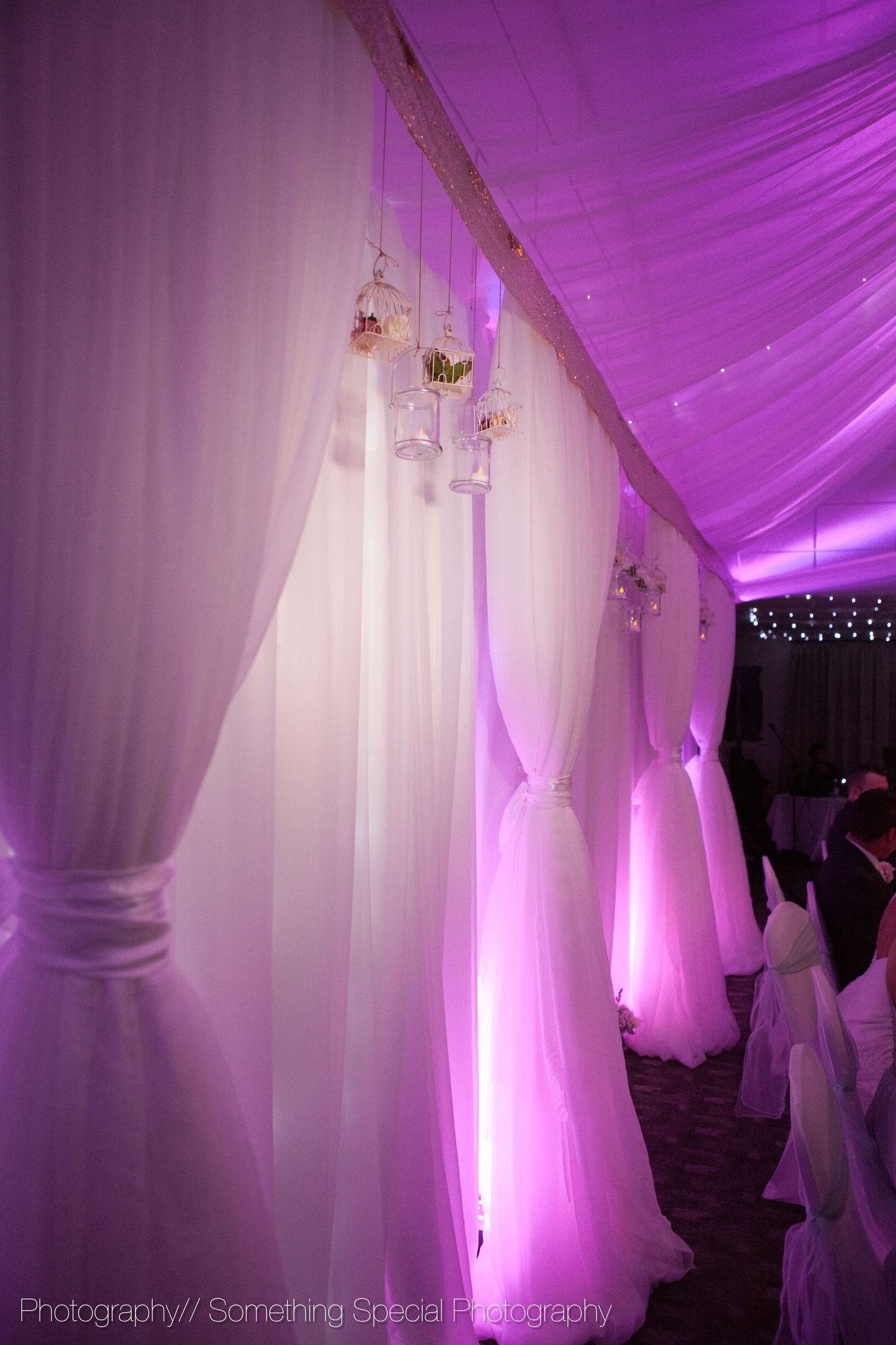 Purple uplighting and white fabric draping create an ethereal wedding reception ambiance with hanging floral accents.