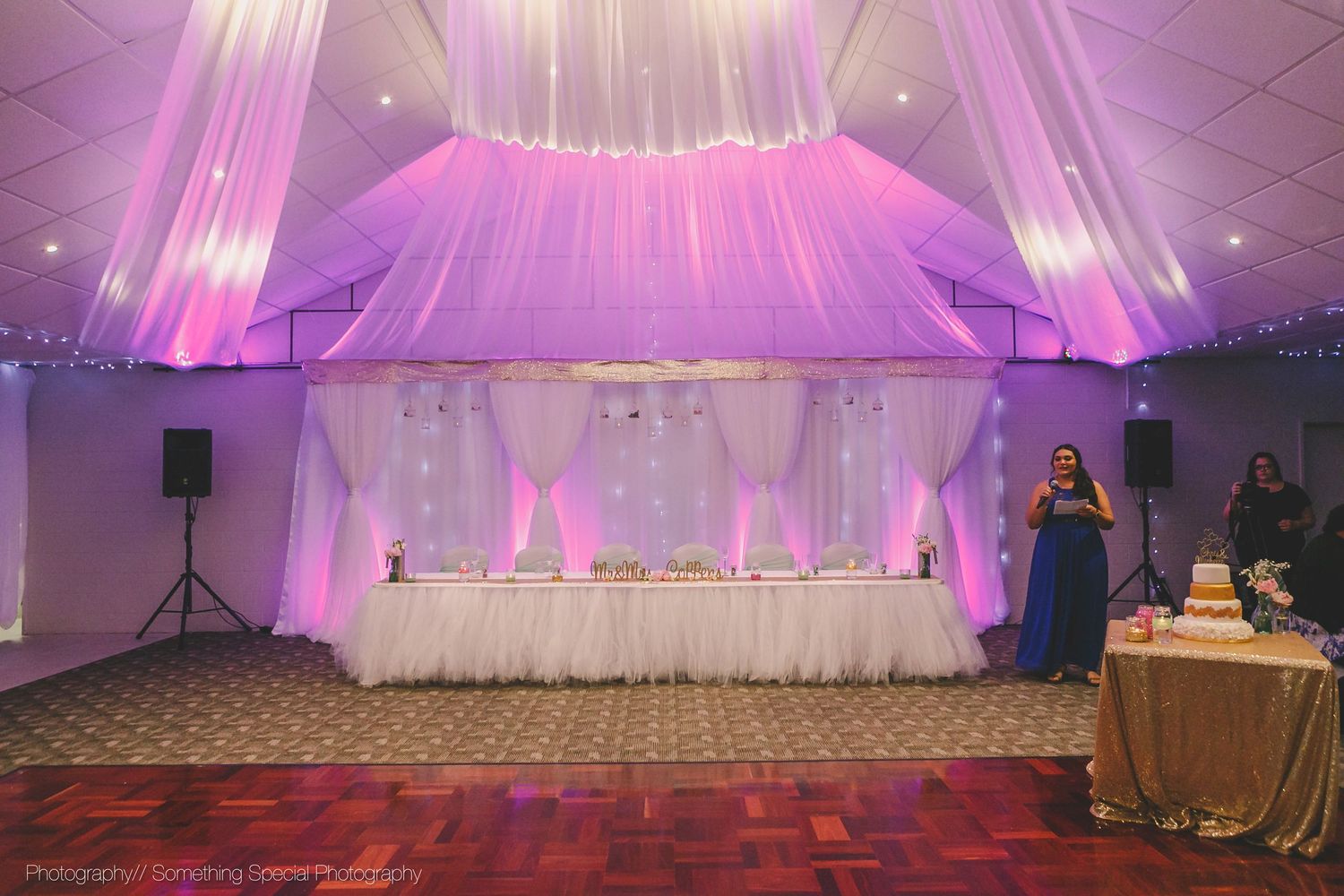 Wedding reception venue decorated in pink and purple lighting with white drapery and head table setup in an elegant ballroom.