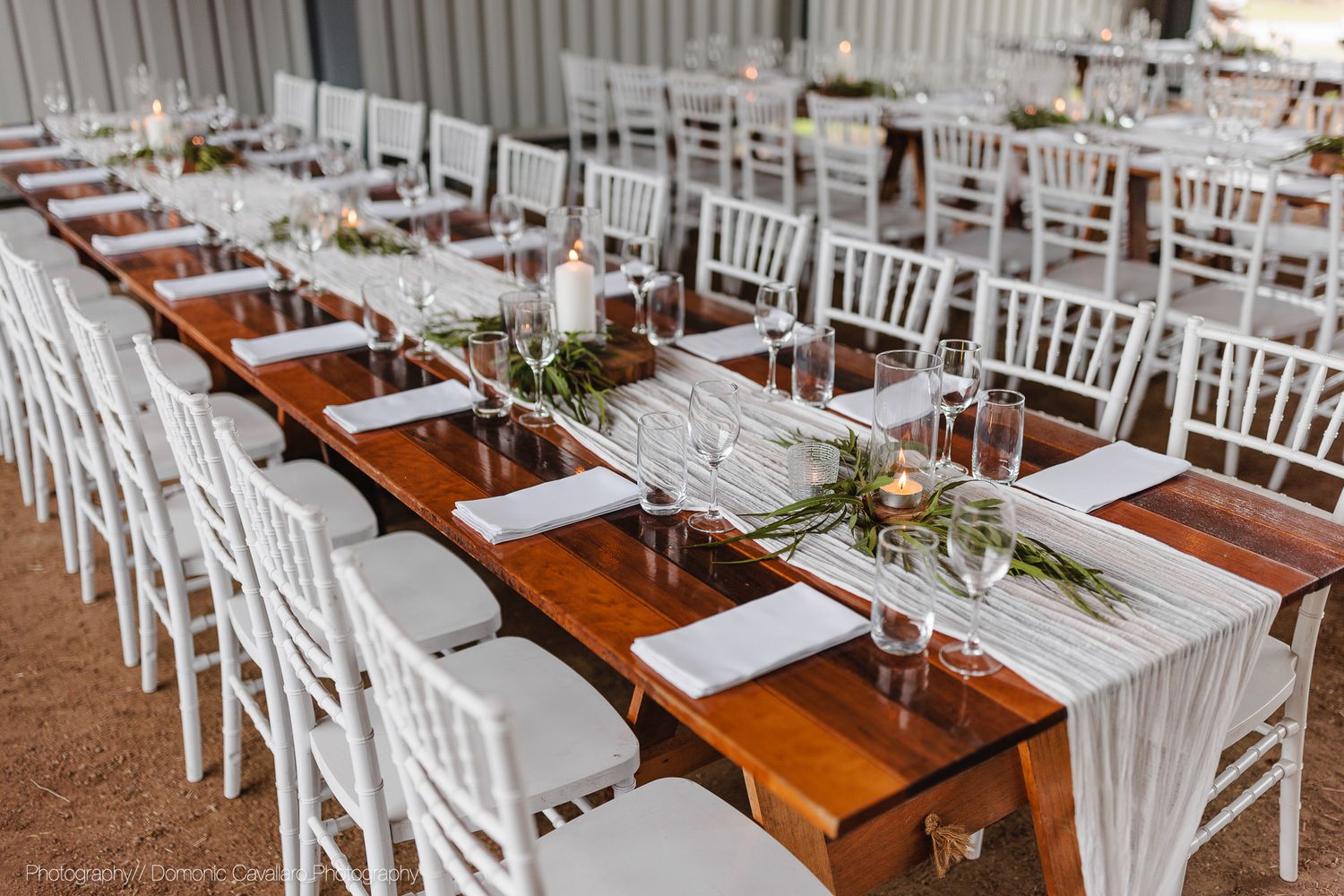 Long wooden tables set for wedding reception with white chairs and greenery centerpieces.