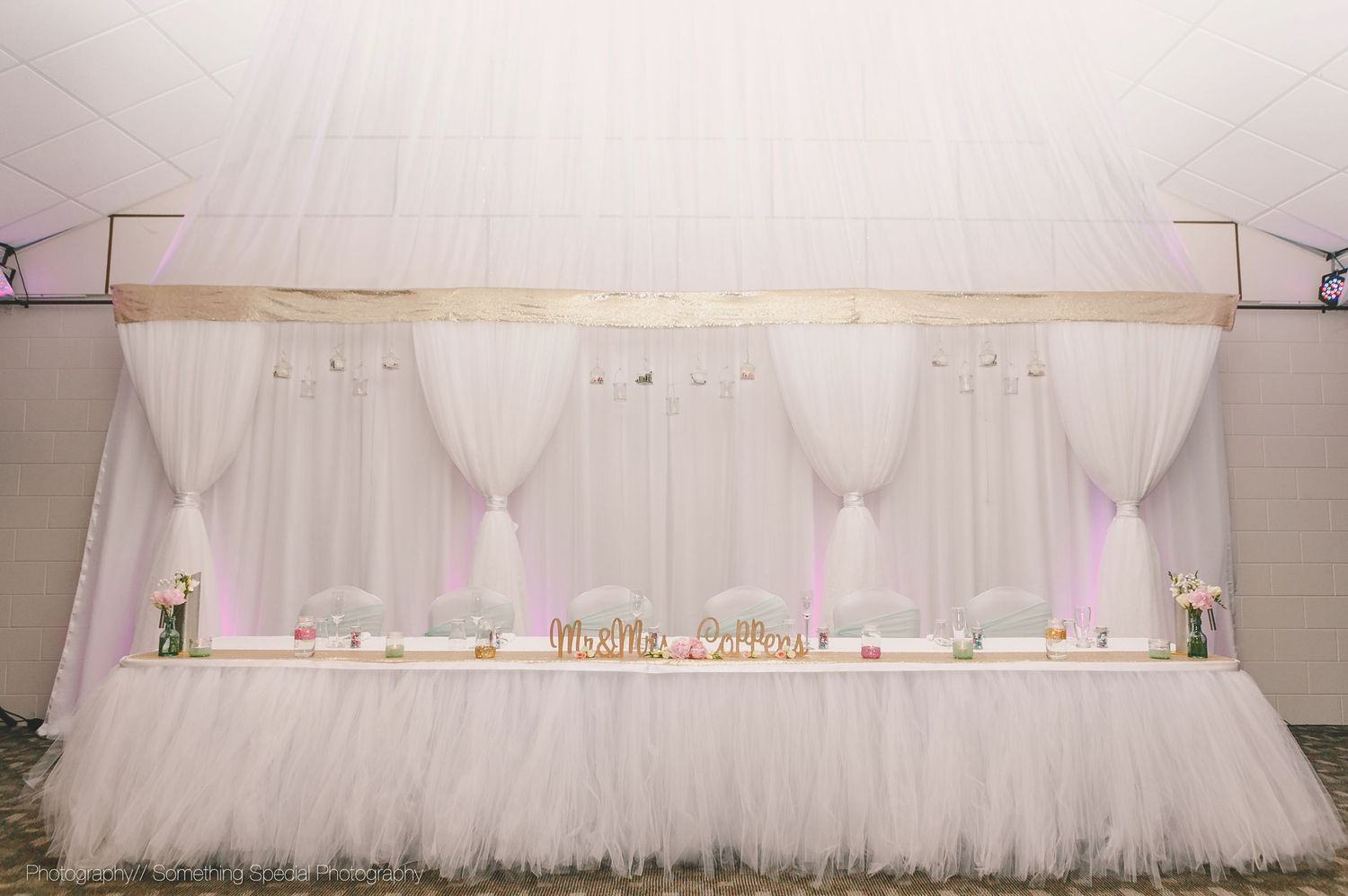 An elegant white tulle skirted head table with sheer draping backdrop and decorative elements at a wedding reception.
