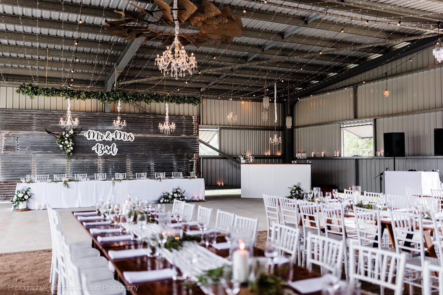 Modern barn wedding reception setup with white chairs, wooden tables and crystal chandeliers.