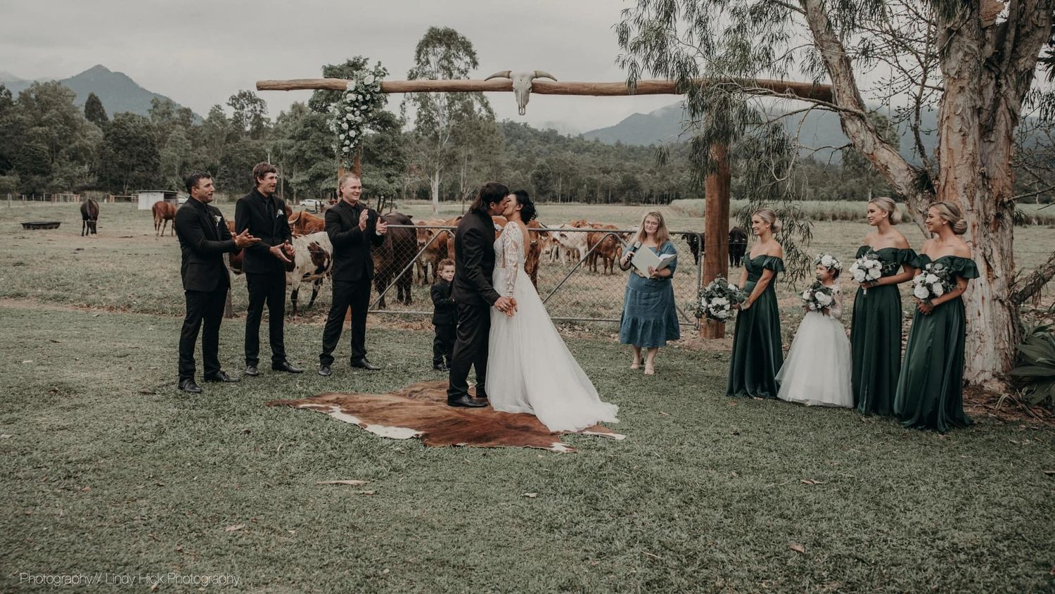 Outdoor wedding ceremony with wedding party and guests gathered on grassy field with mountains and eucalyptus trees in background.