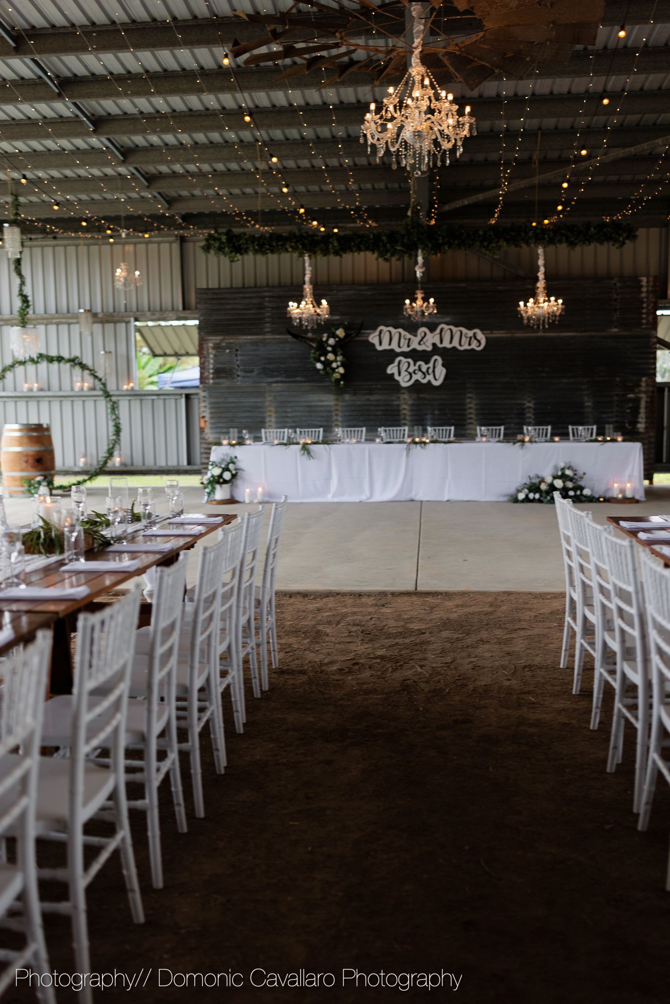 Rustic wedding reception venue with chandeliers, wooden tables, and white chairs arranged in rows.
