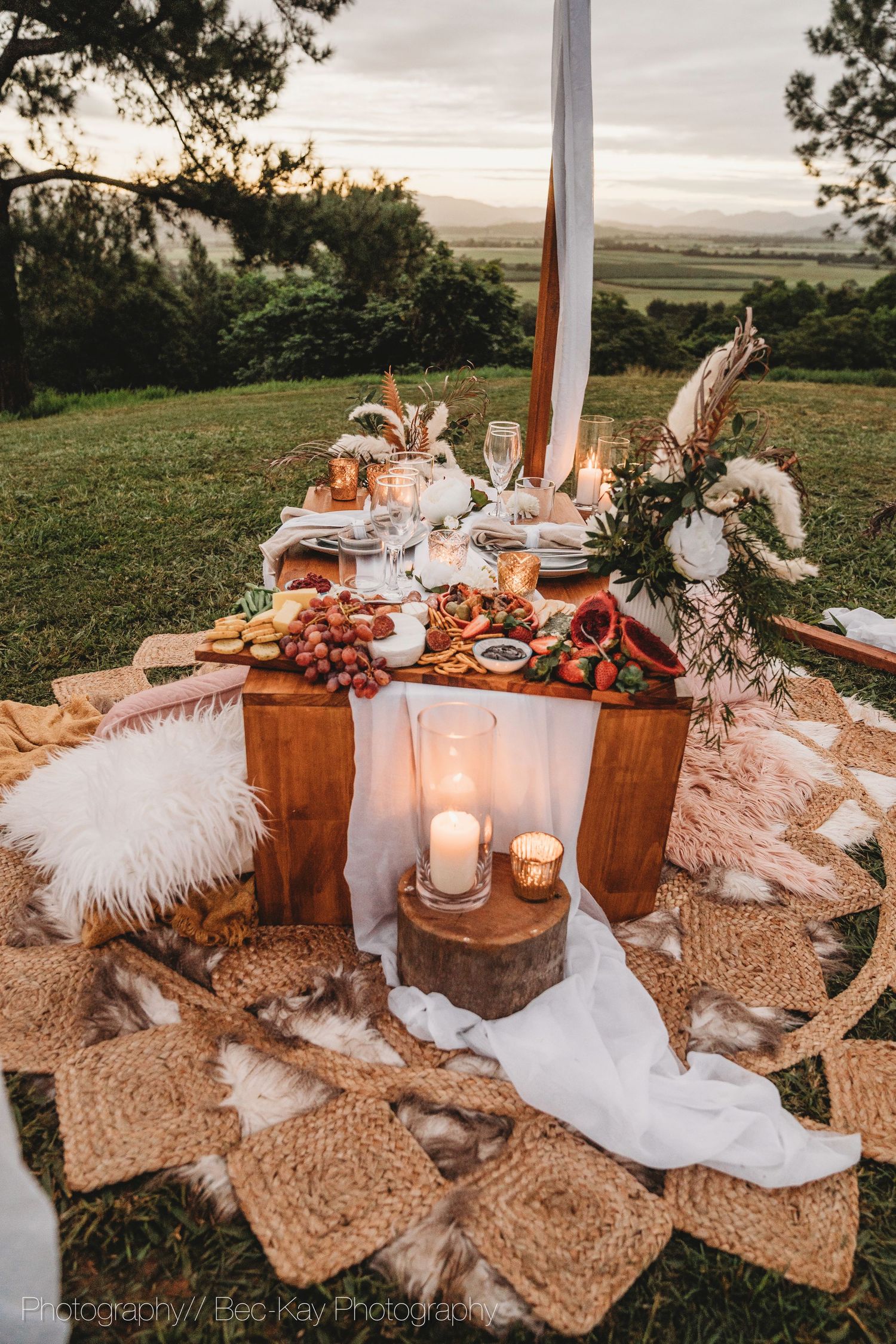 Rustic outdoor picnic setup with white linens, candles, fur cushions and grazing board overlooking scenic valley view.