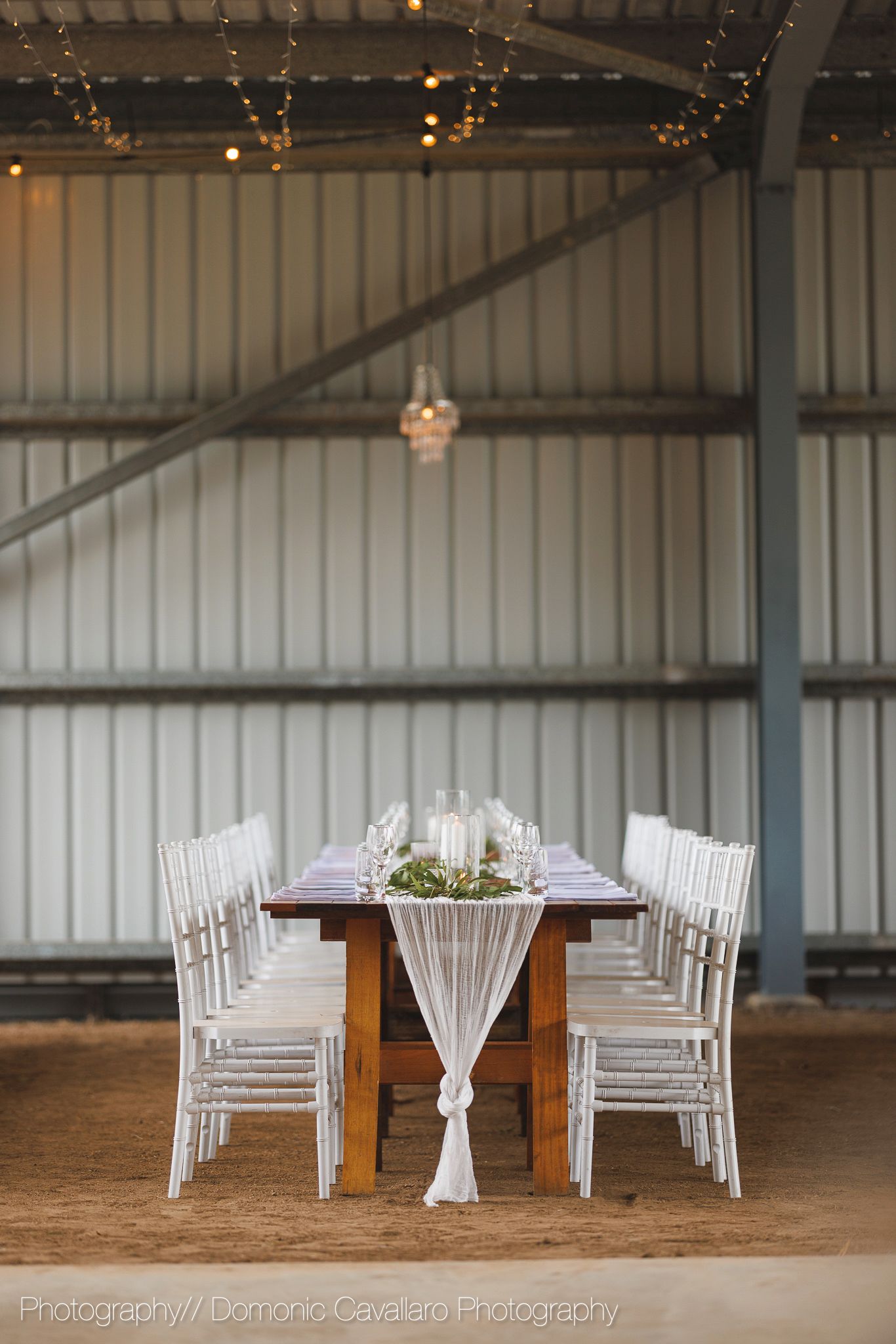 Long wooden dining table with white chairs and elegant table settings in an industrial-style venue.