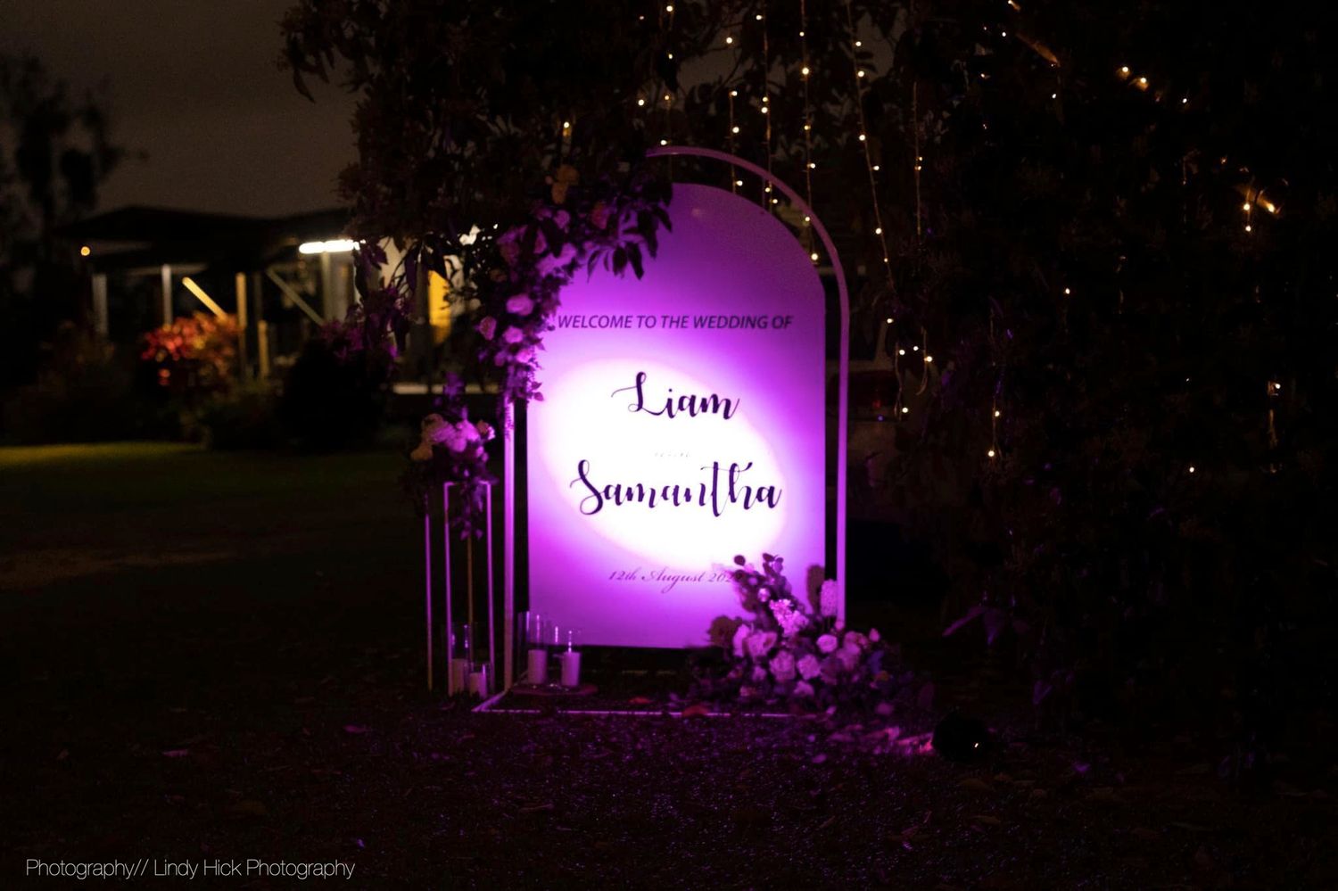 Illuminated wedding sign with purple backlight and floral decorations at night.