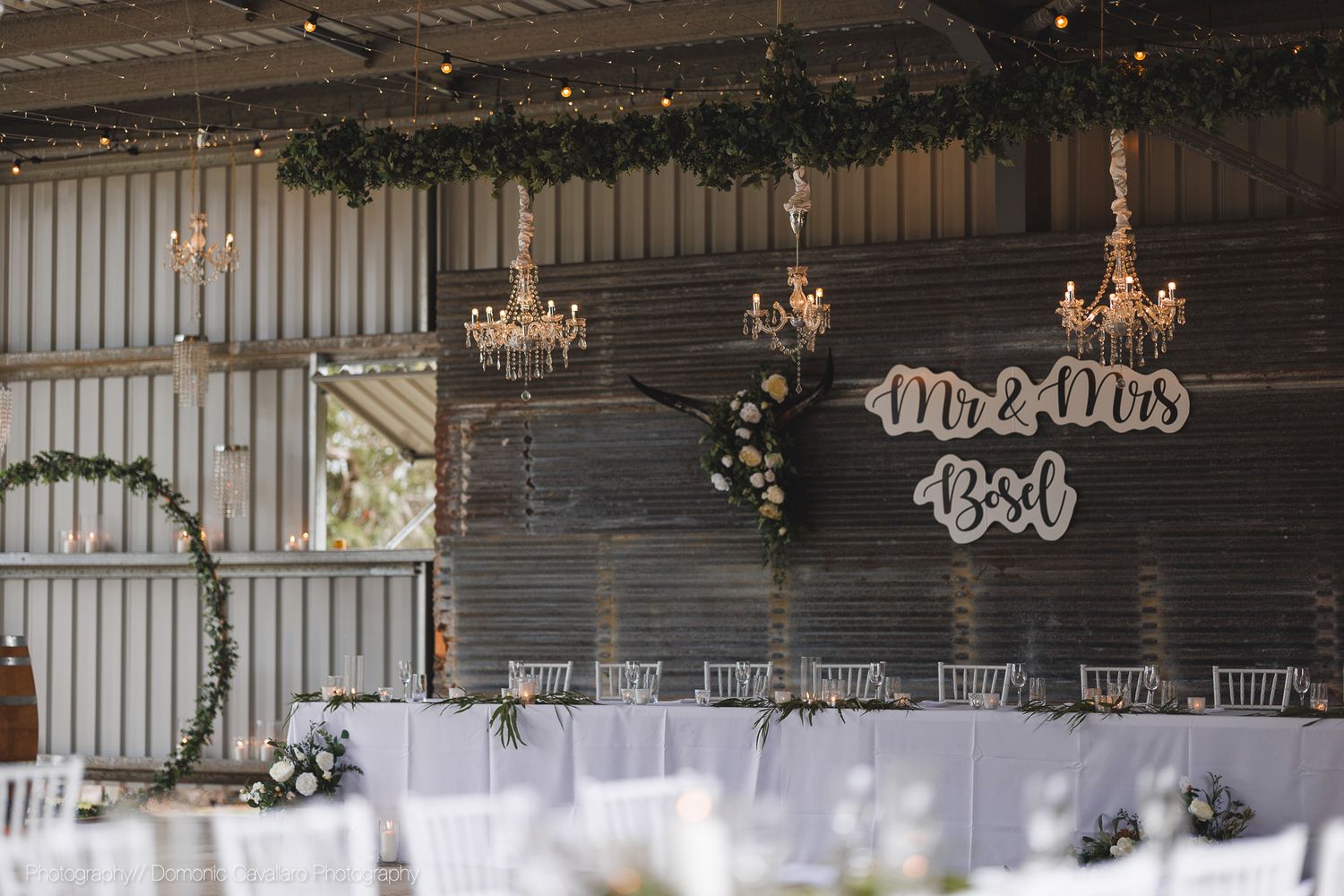 Elegant barn wedding reception setup with crystal chandeliers and 'Mr & Mrs' wall sign in rustic venue.