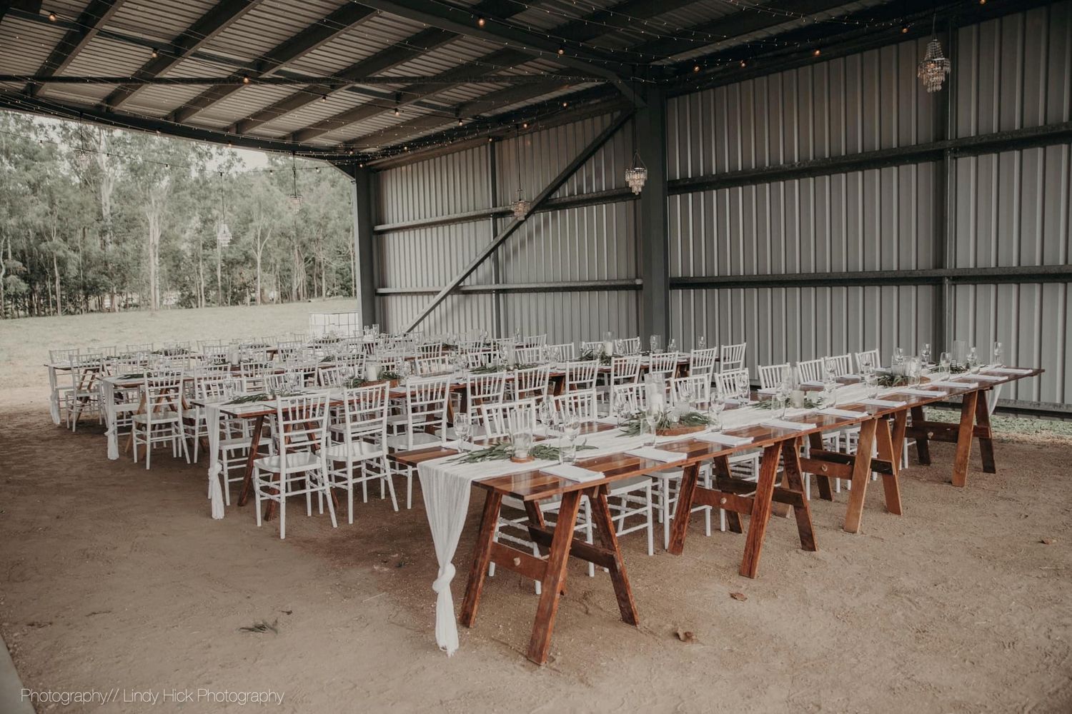 White wooden chairs and rustic tables are arranged in rows inside an industrial barn-style wedding venue setting.