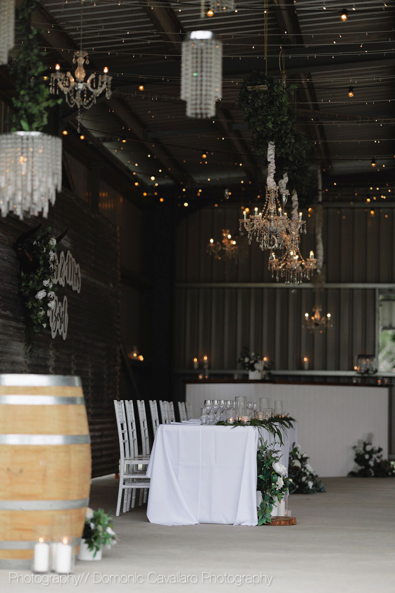 Wedding venue interior with crystal chandeliers, wine barrel, and white table settings in romantic lighting.