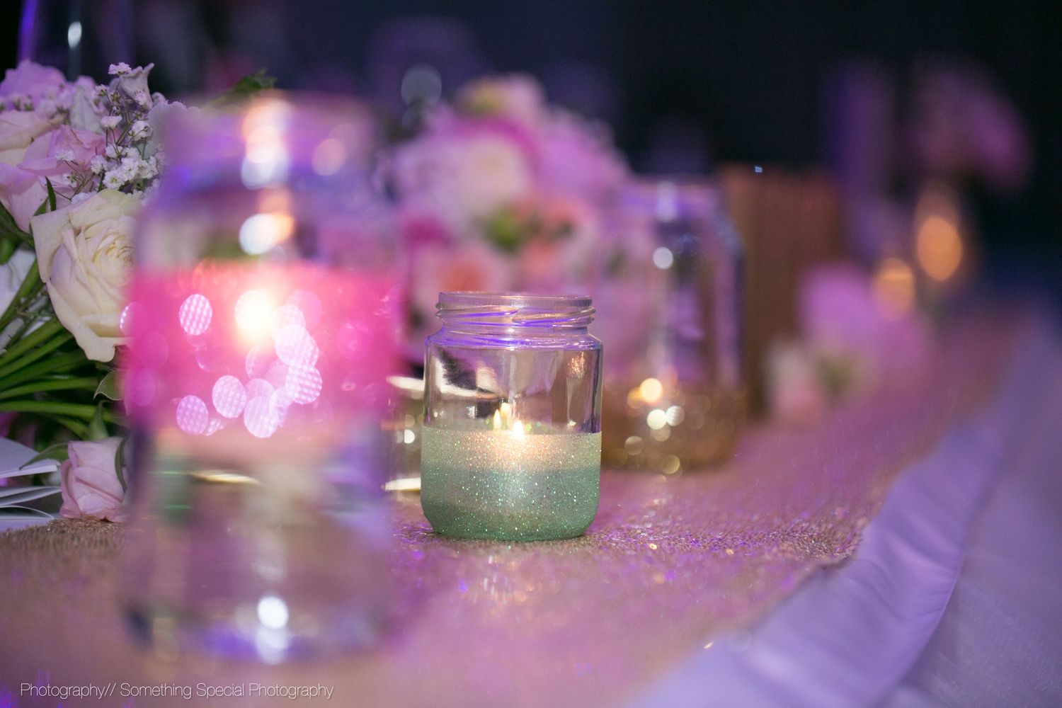 Small mason jar candle glowing with pink and purple hues on a decorated table with floral decorations and soft lighting.