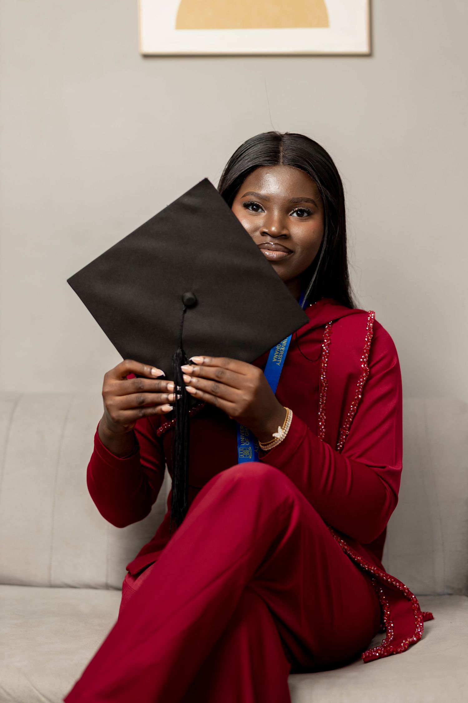 Young woman in red suit holding black graduation cap while sitting on a couch, celebrating academic achievement.