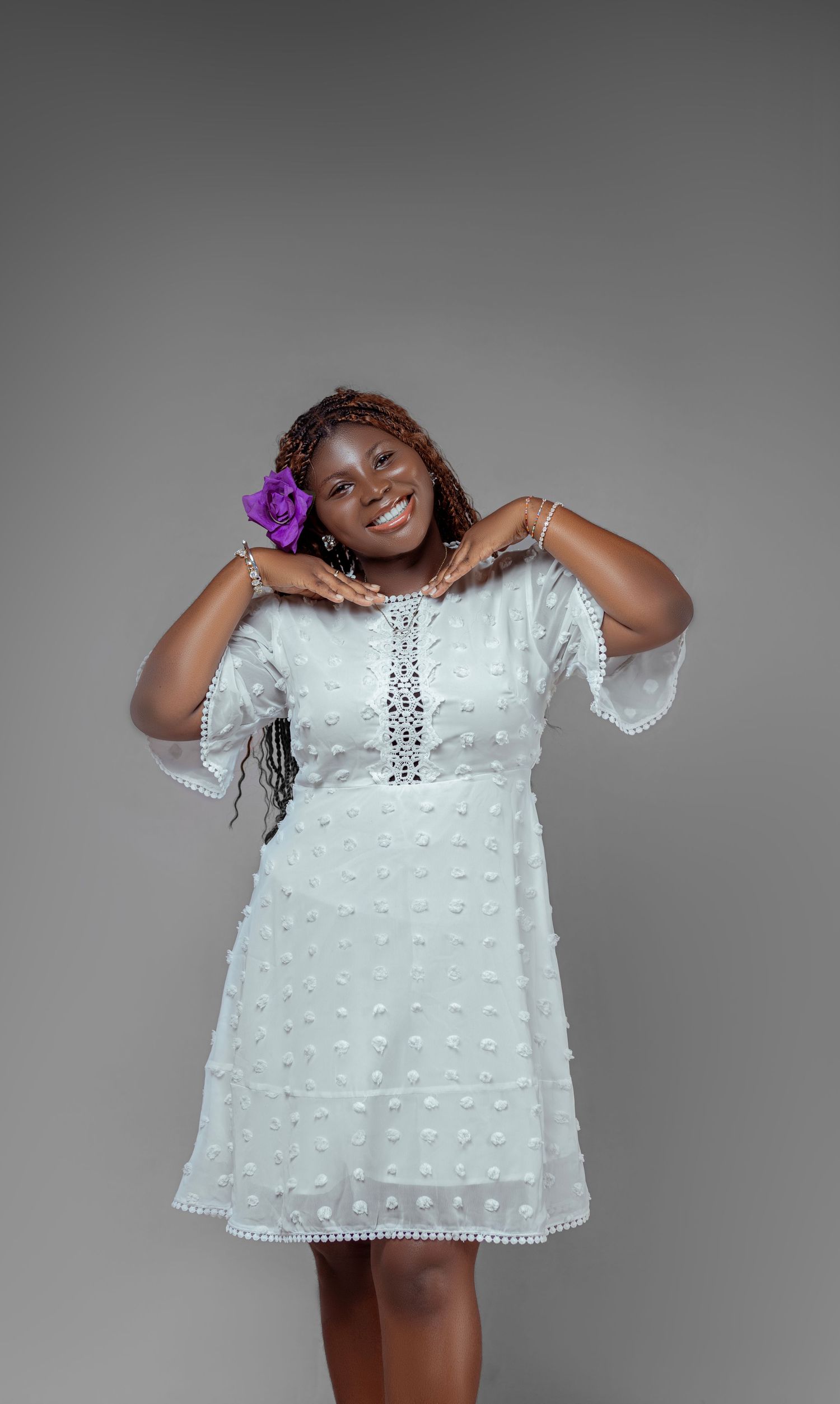 A woman in a white dotted dress poses elegantly with a purple flower in her hair against grey background.