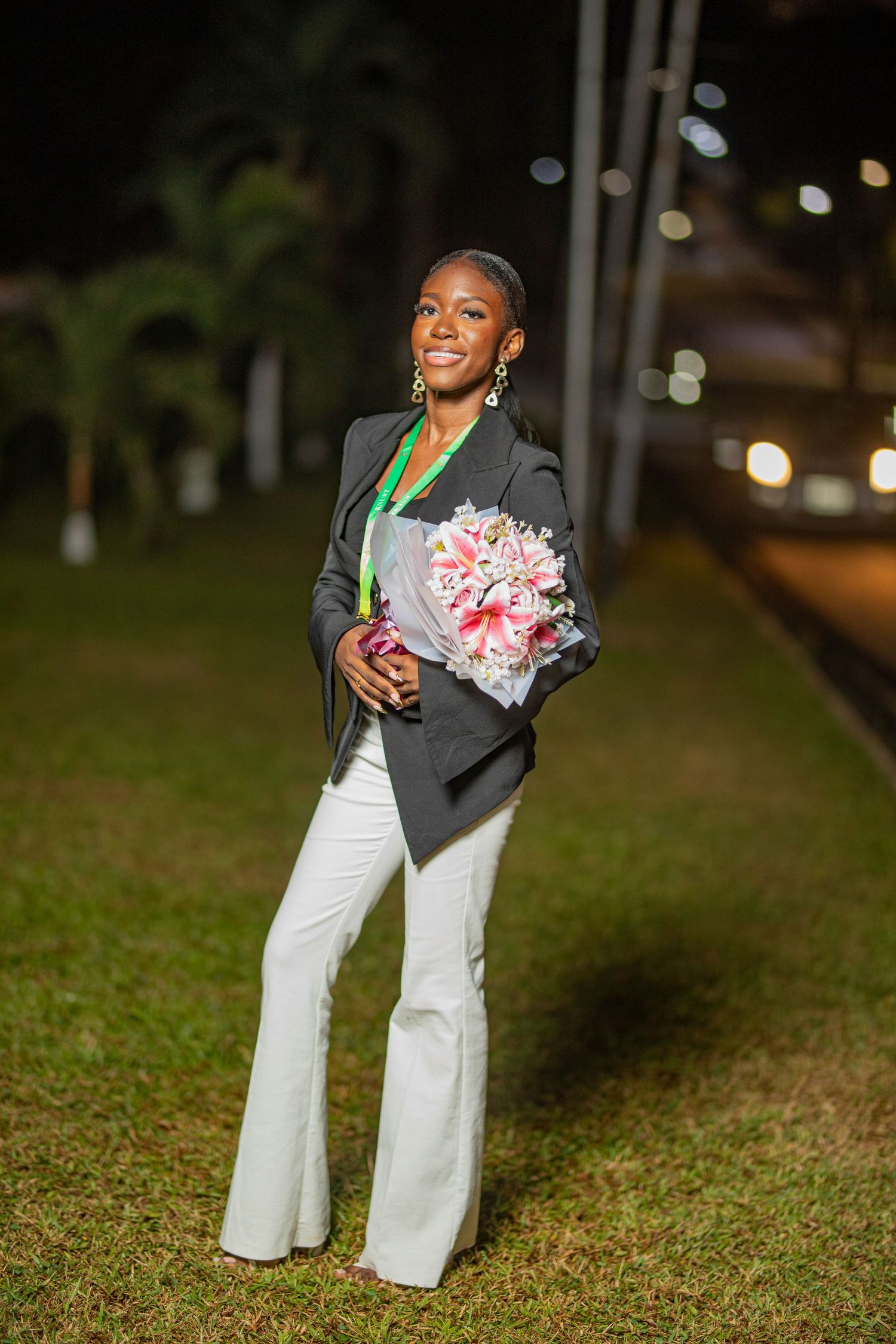 A person in a gray blazer and white pants poses joyfully on grass while holding pink and white flowers.