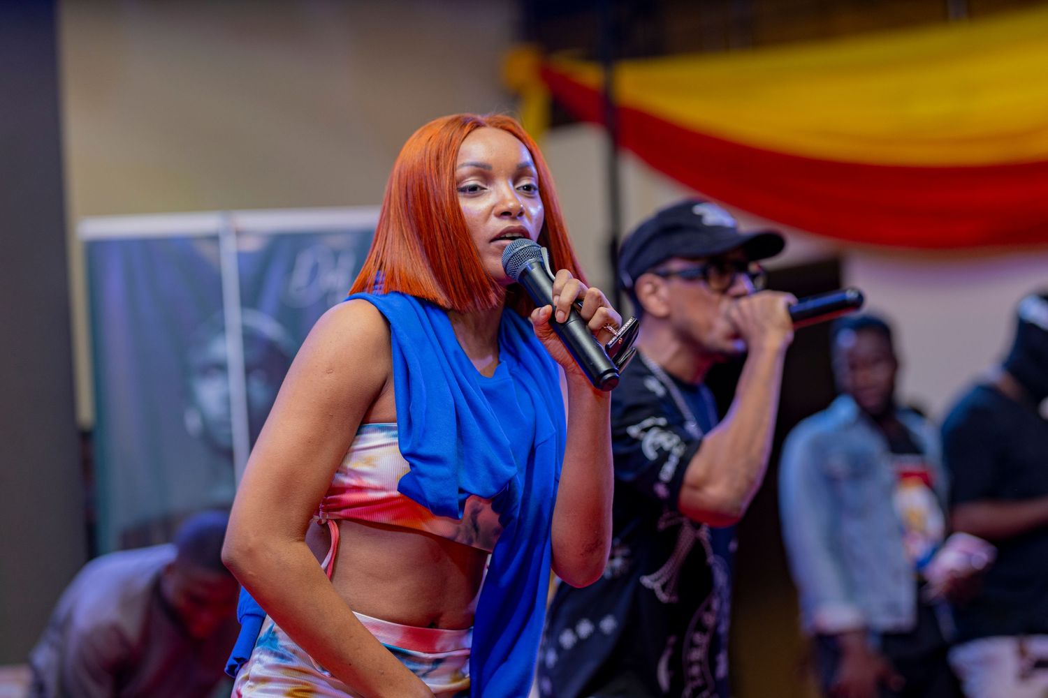 Performer in blue crop top sings passionately on stage with colorful backdrop.