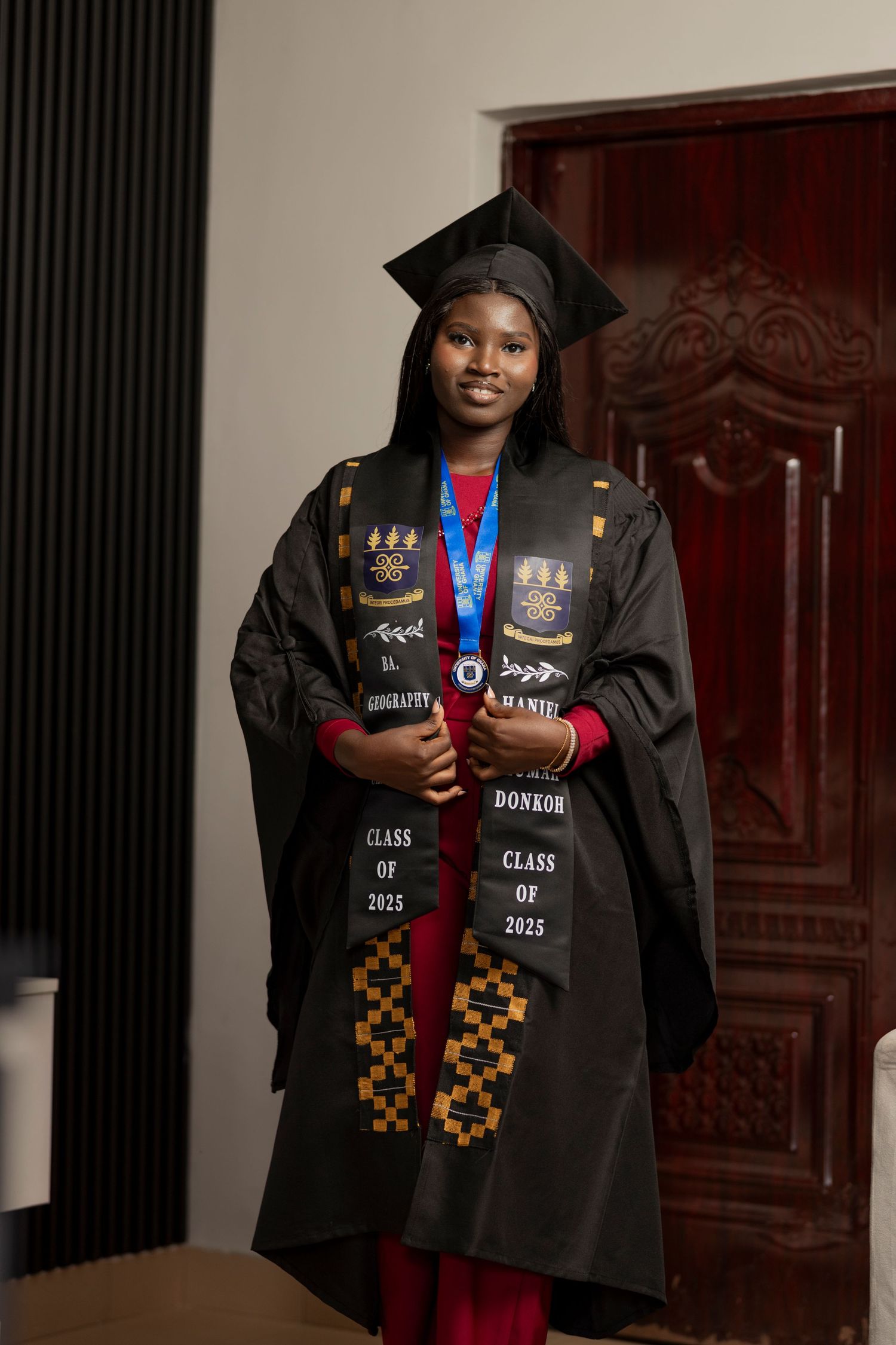 Young woman in black graduation gown and cap wearing Class of 2025 stoles and medal, smiling indoors.