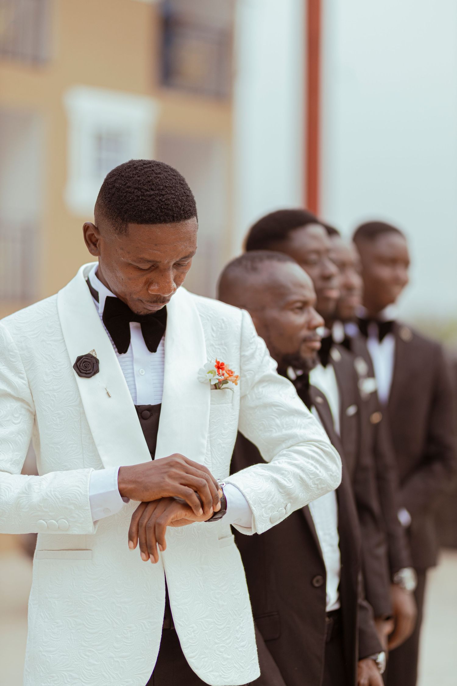 Groomsmen in formal attire with the groom wearing a white tuxedo jacket checking his watch.