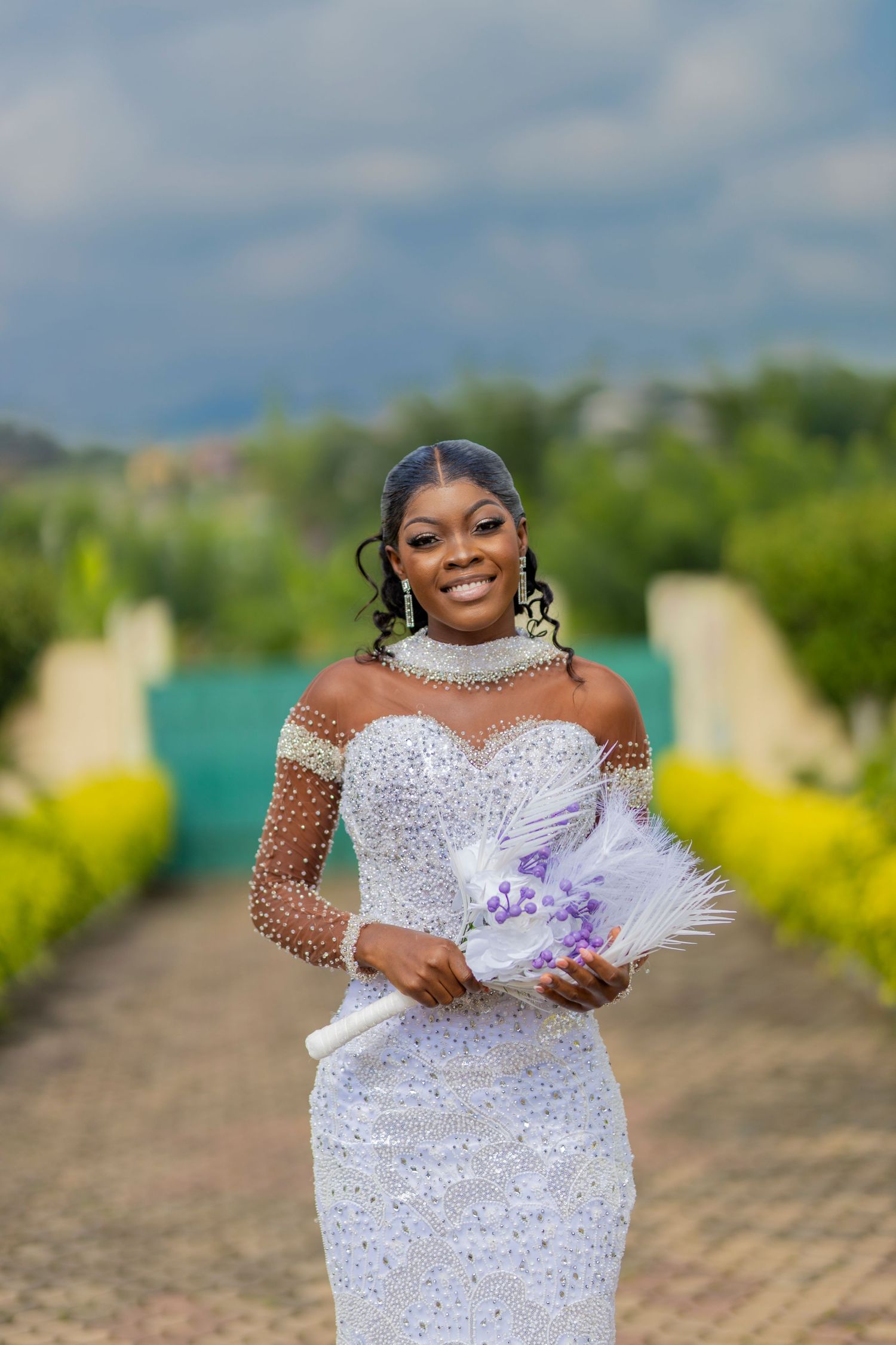 A stunning bridal portrait in a white beaded gown with feather fan accessory against a blurred outdoor garden backdrop.