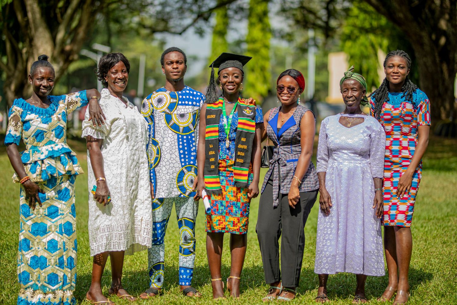 A group of people wearing colorful African print clothing and formal attire pose together outdoors on grass.