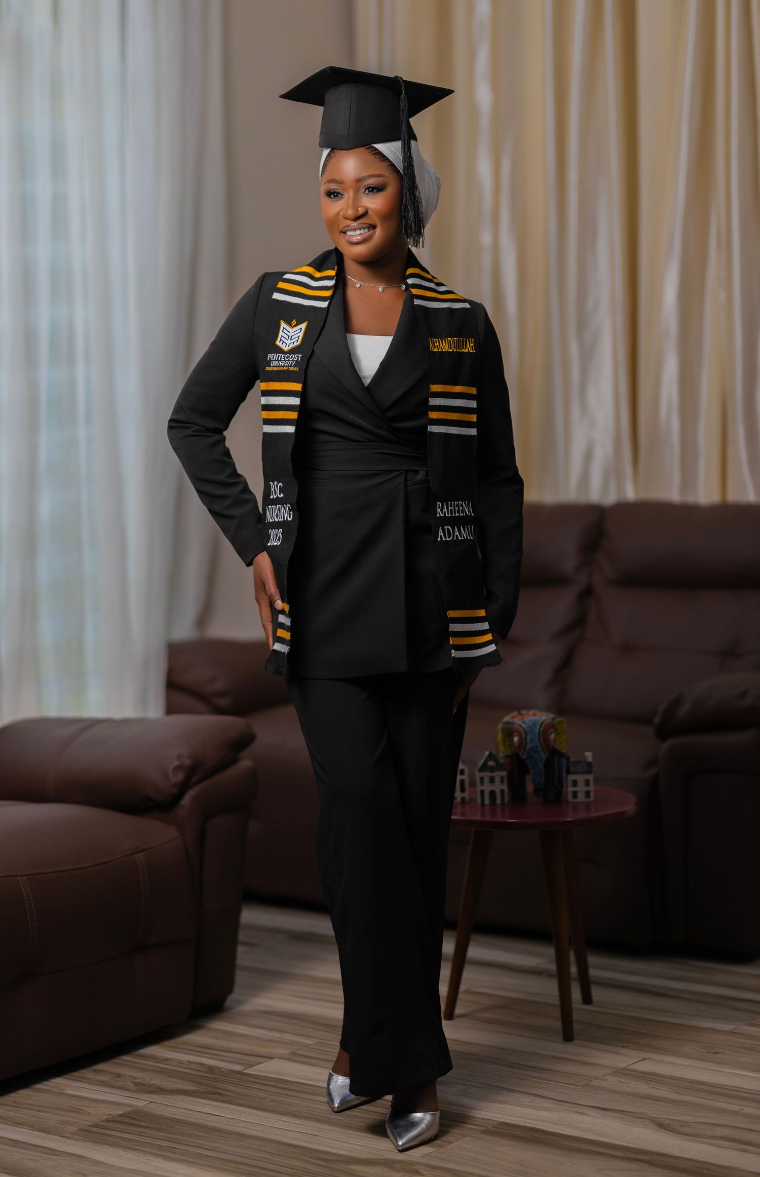 Graduate in black suit and graduation cap wearing a decorative stole stands proudly in living room setting.