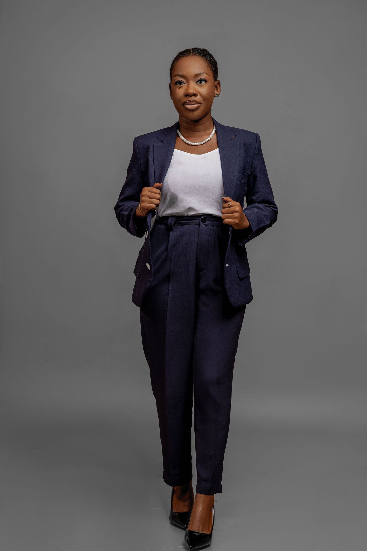 Confident professional wearing navy business suit with white top and pearl necklace poses against gray backdrop.