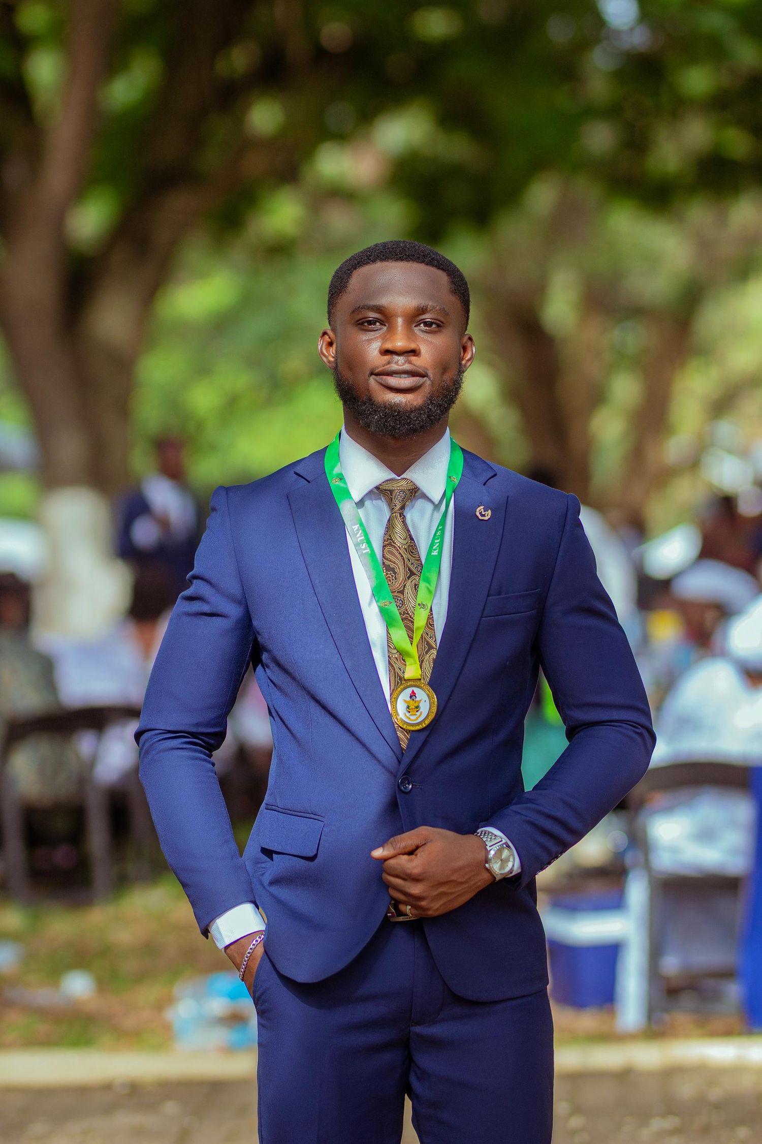 Graduate in navy blue suit poses with academic medal during outdoor commencement ceremony.