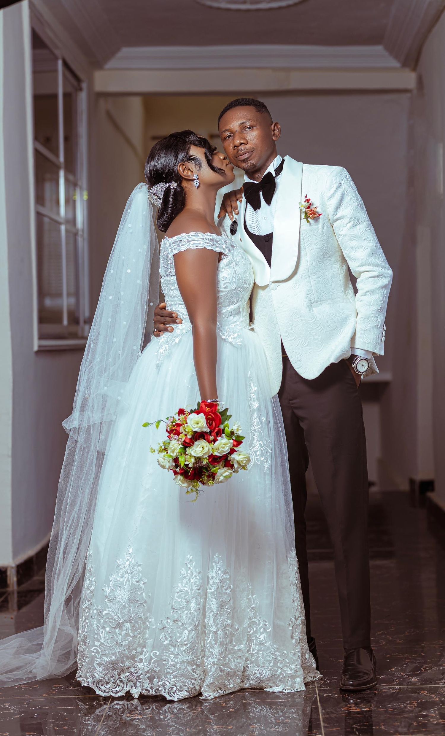 Newlyweds in elegant white wedding attire pose with red bouquet in indoor corridor.