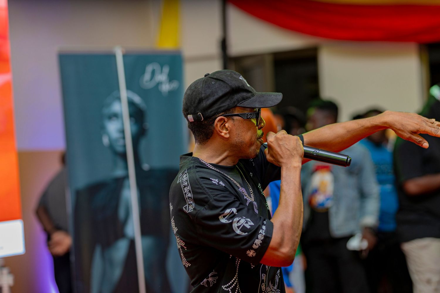 Performer in black attire and cap energetically raps on stage at outdoor event.