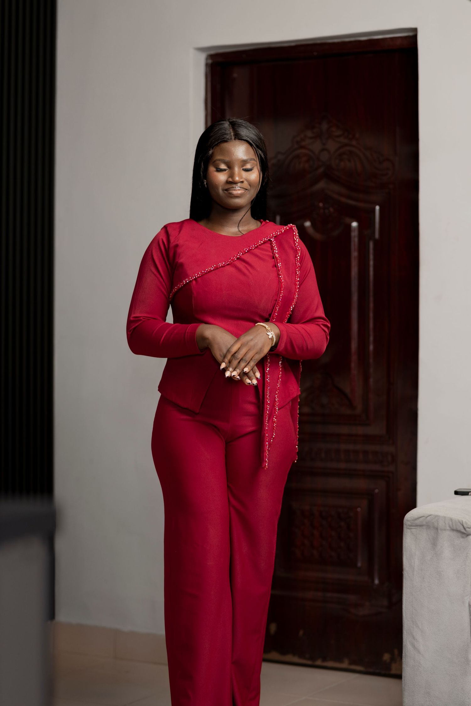 Woman in elegant red long-sleeve top and wide-leg pants standing gracefully indoors near a wooden door.