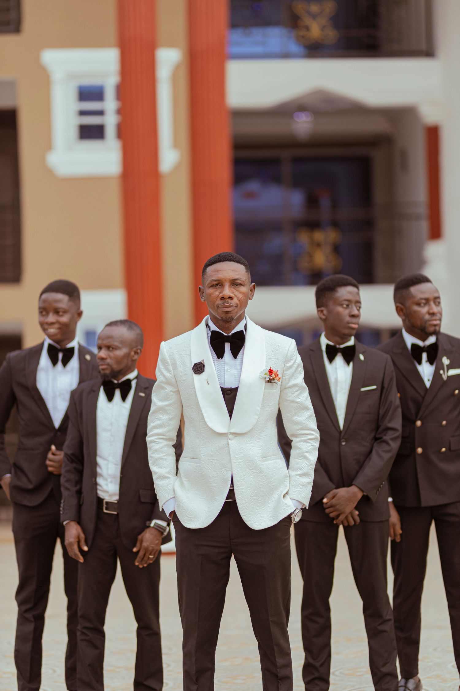 Wedding party lineup featuring groomsmen in dark suits and white tuxedo jacket in the center.