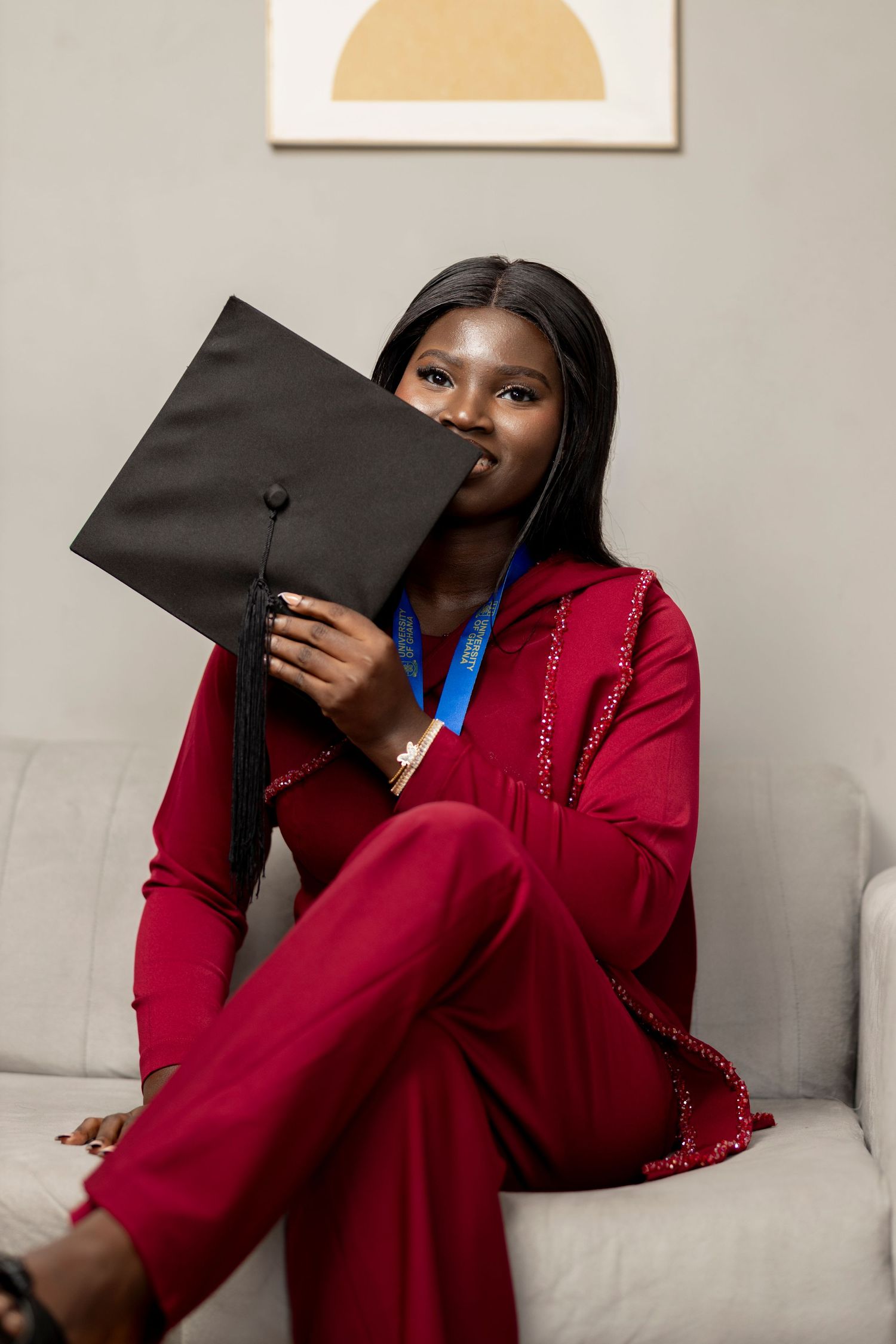 A young Black woman in a red outfit holds a black graduation cap while sitting on a gray sofa, smiling proudly.