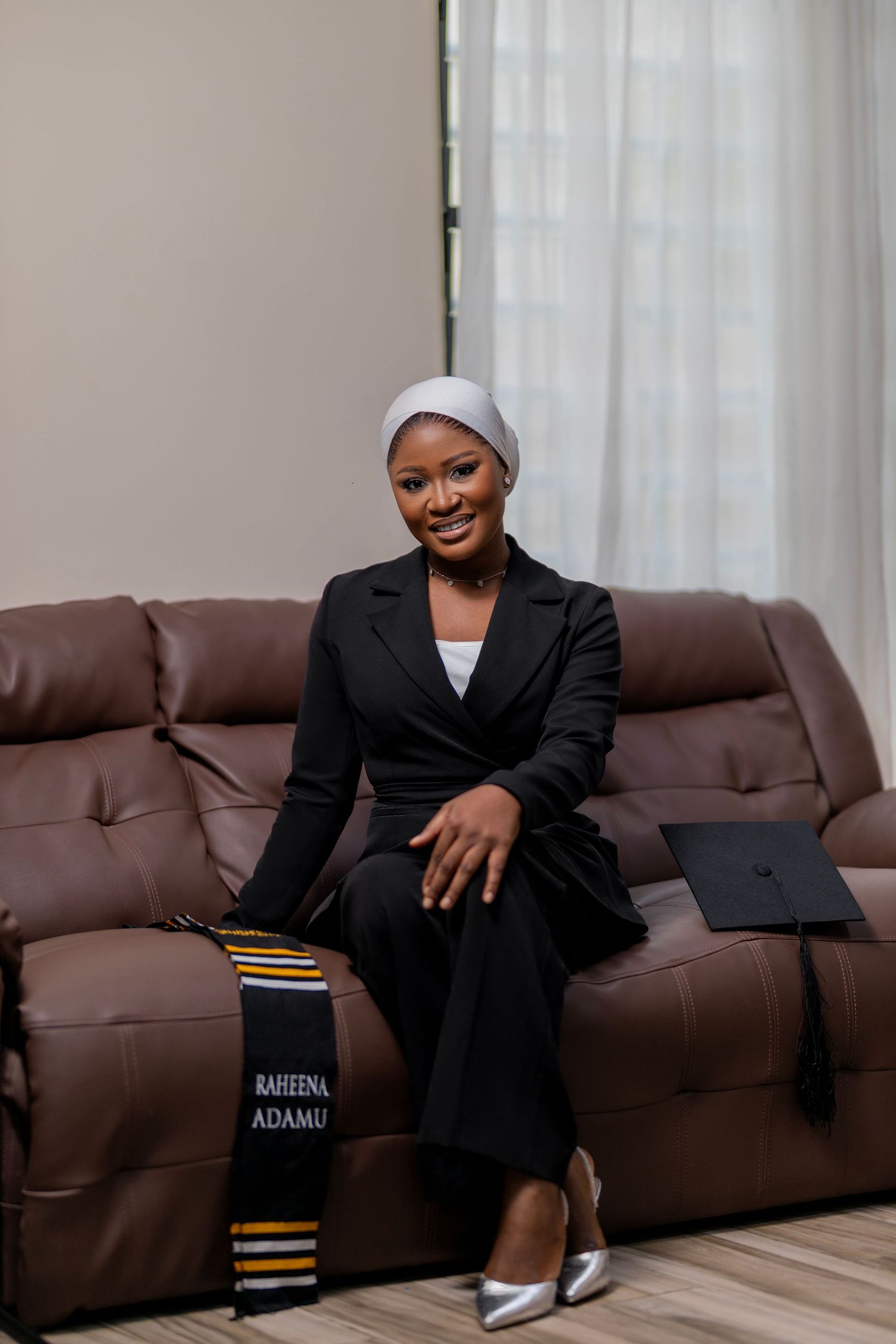 Graduate in black suit sits on brown couch with graduation cap and stole nearby.